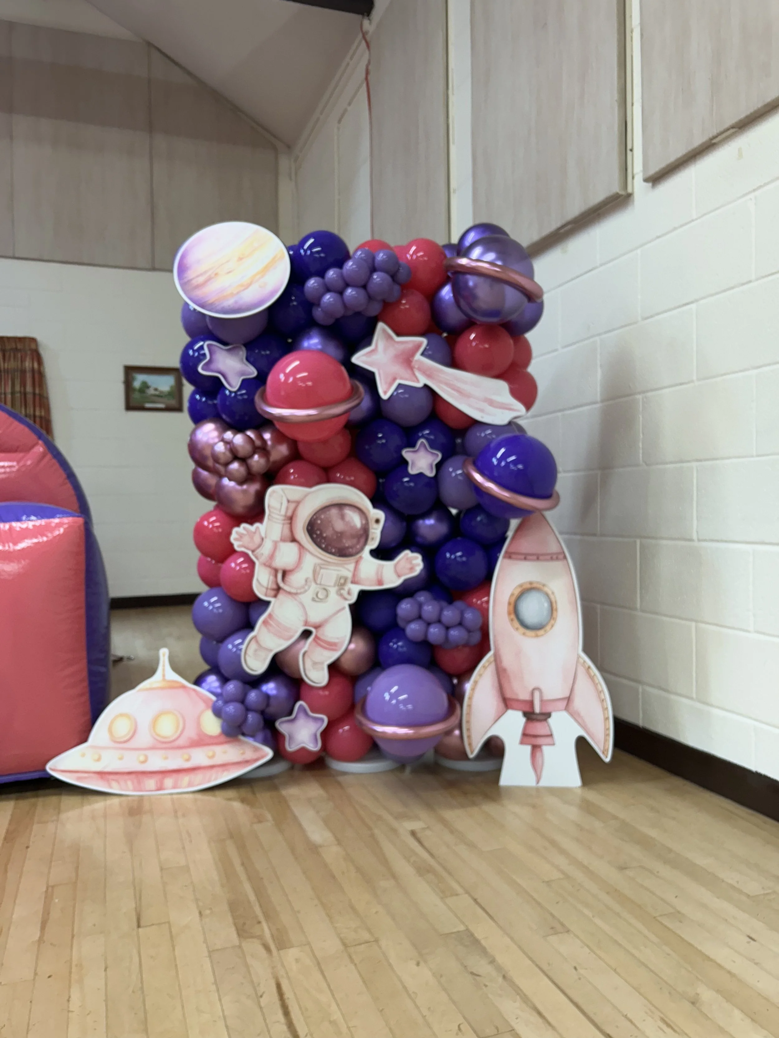 Decorative space-themed balloons with planets, stars, a rocket, and an astronaut cutout arranged on a stand inside a room with wooden flooring and paneled walls.