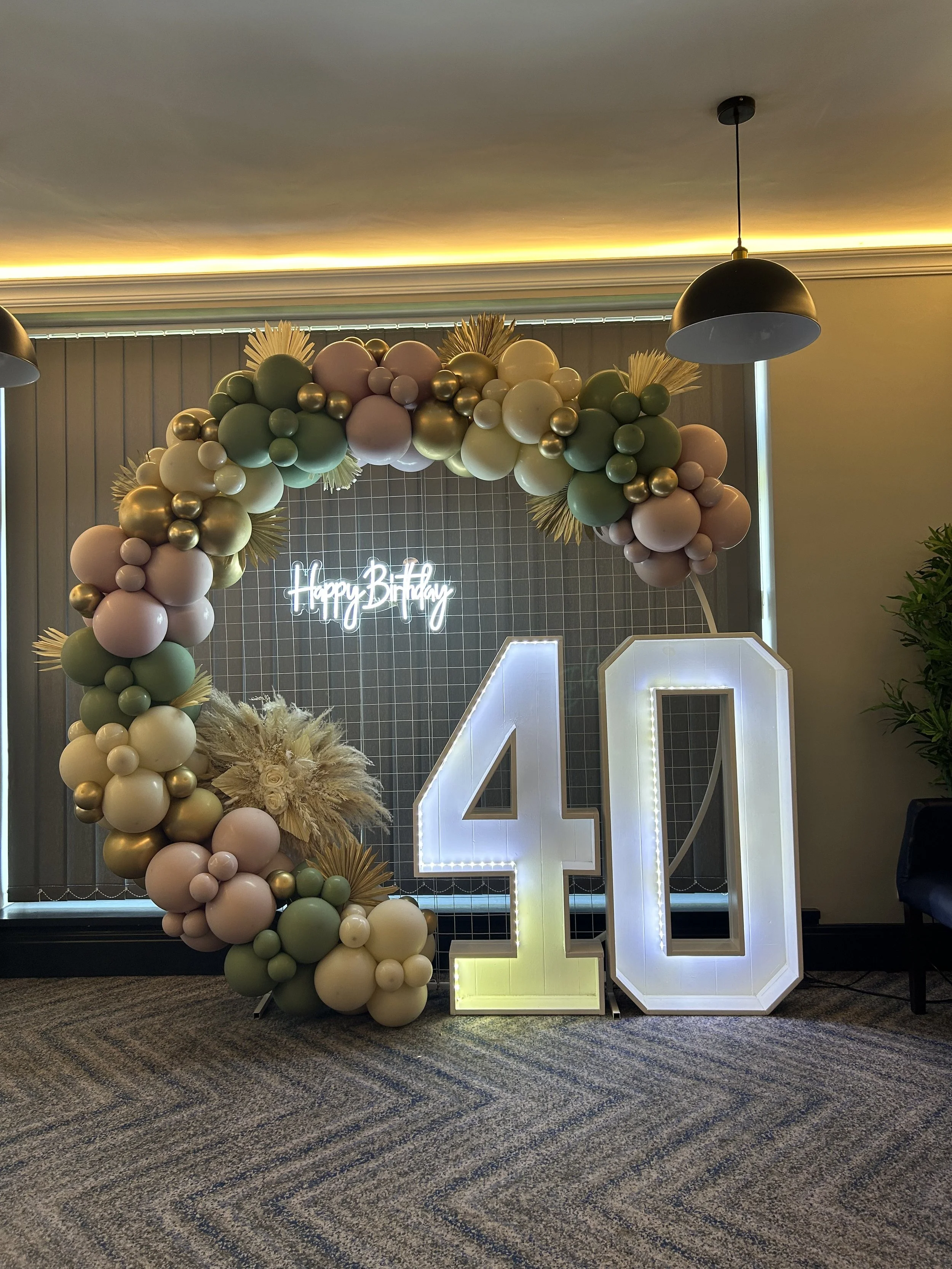 Decorative balloon arch with pink, green, cream, and gold balloons, surrounding a large illuminated number 40 and a neon 'Happy Birthday' sign in the background.