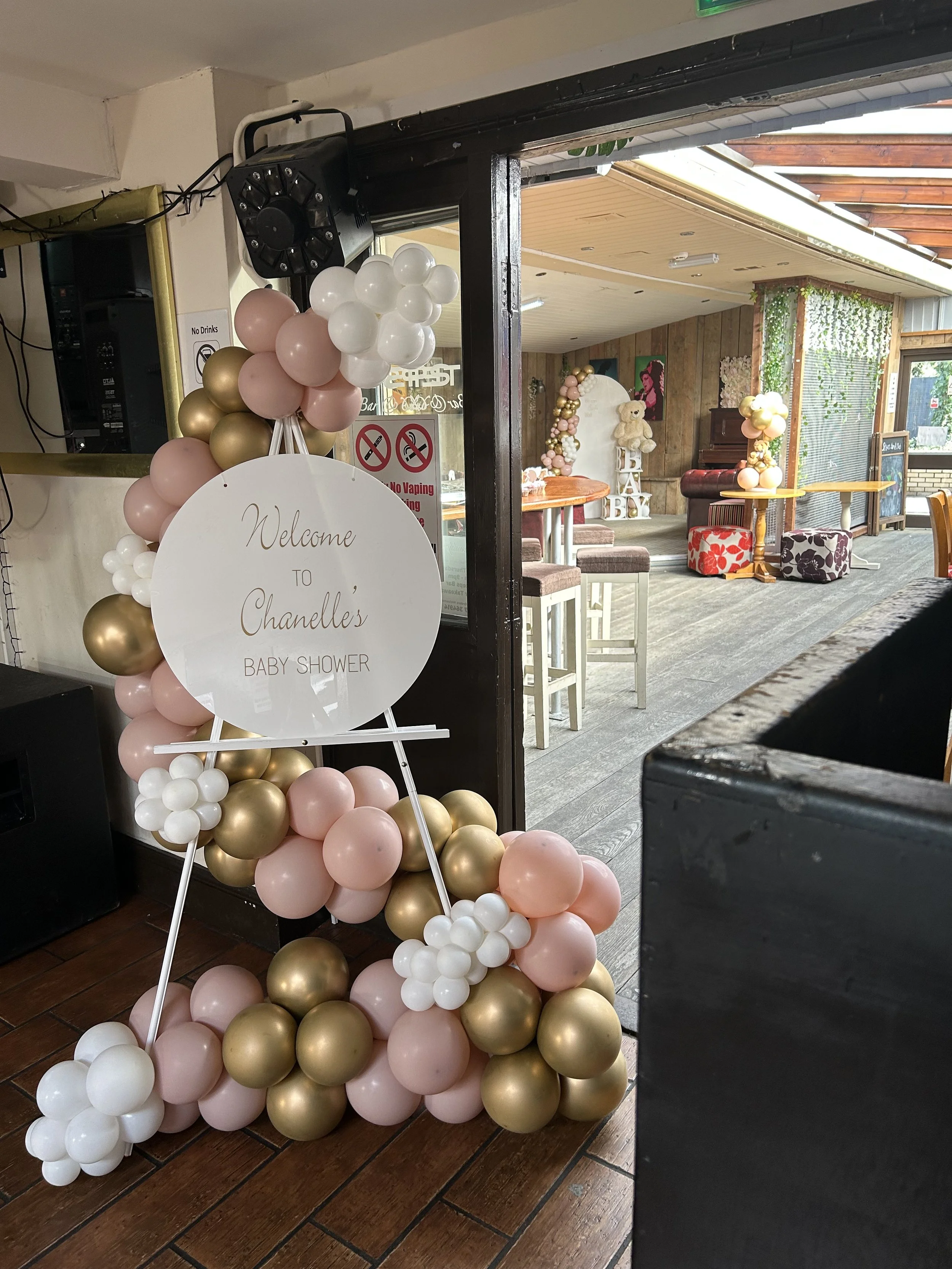 Decorative balloon arrangement with pink, white, and gold balloons, and a welcome sign for Chanelle's baby shower inside a festively decorated room with a teddy bear and flower wall art.