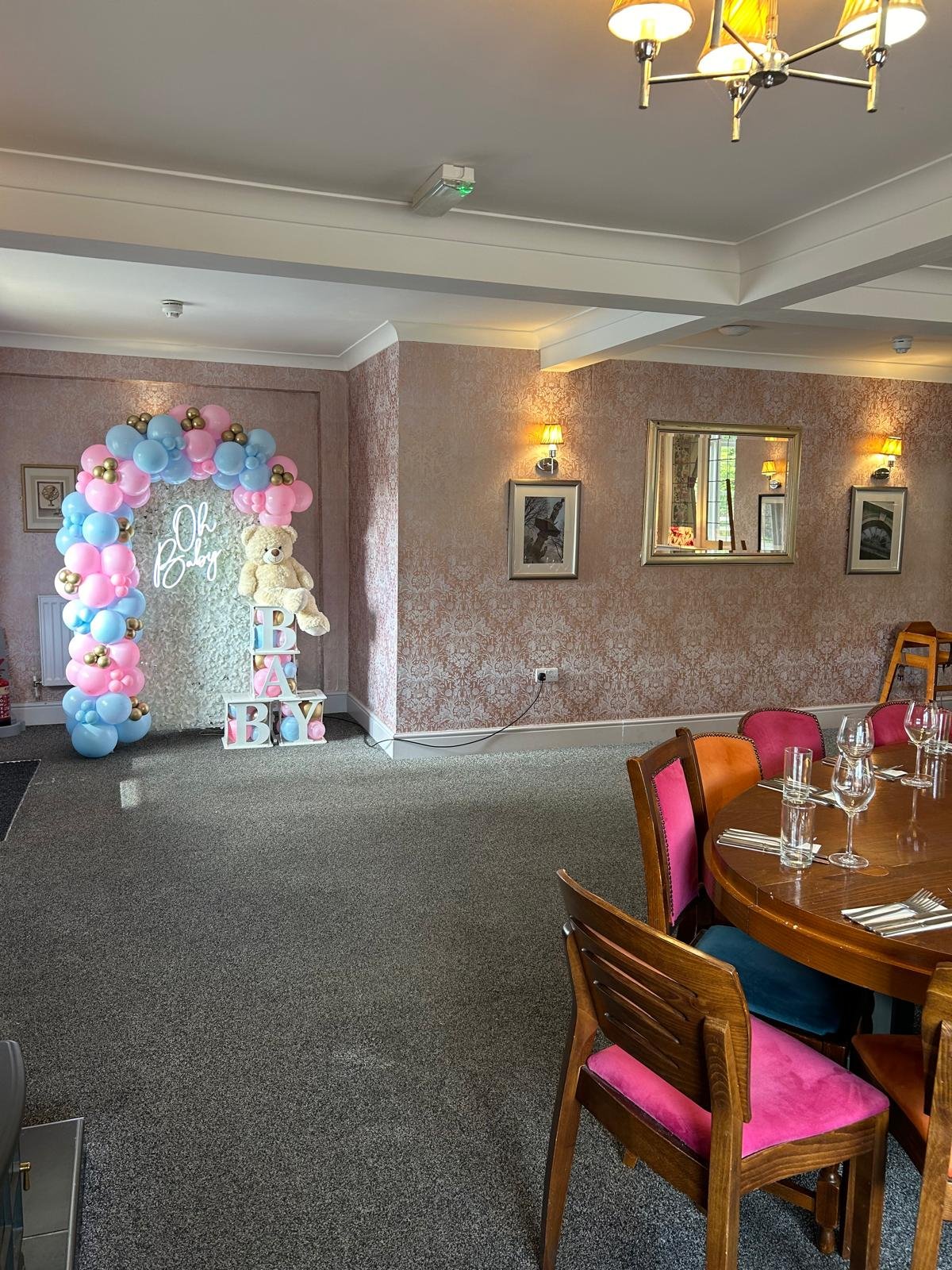 A decorated indoor space for a baby shower, with a balloon arch, a teddy bear, and blocks spelling 'BABY' in front of a white textured backdrop, with the words 'Oh Baby' written in neon lights. The room has a carpeted floor, pink and orange upholstered chairs around a wooden table set with glasses and cutlery, framed pictures on the walls, and warm lighting.