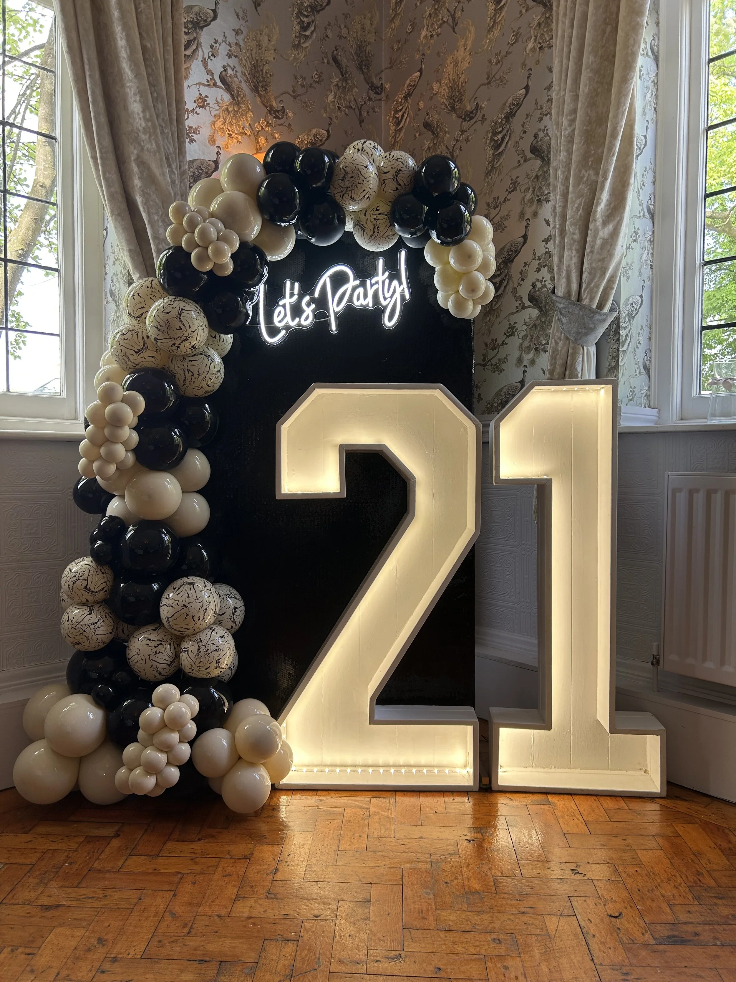 Decorative balloon arch with black, white, and marbled balloons, illuminated white 'Let's Party' sign, and large illuminated number 2 and 1 for a celebration or birthday party inside a room with wooden flooring and patterned wallpaper sign with peacock motifs, flanked by cream curtains.