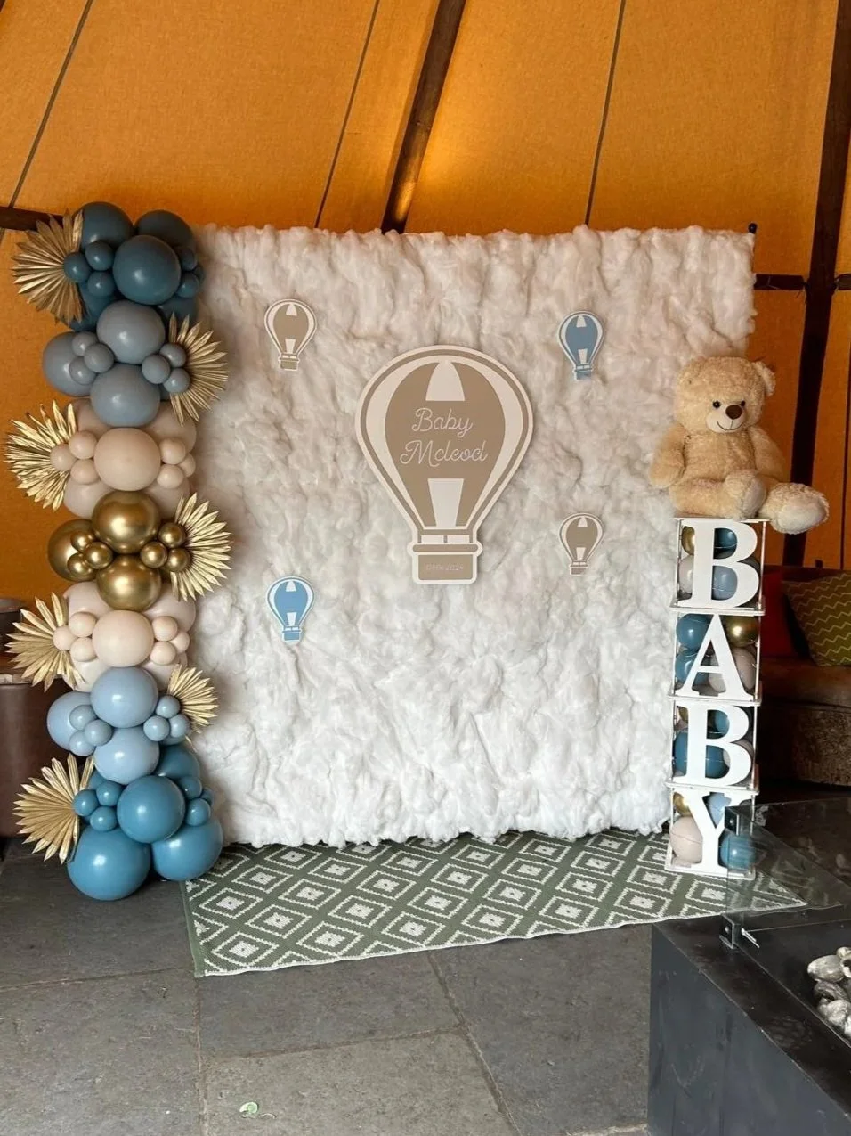 Baby shower decoration with a white fluffy backdrop, blue and gold balloons, a teddy bear, and a sign that reads 'Baby McLeod' inside a hot air balloon design.