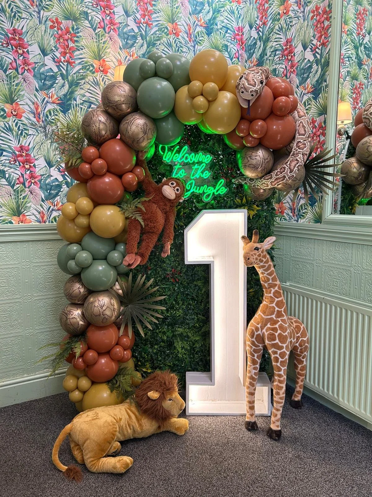 Decorative jungle-themed backdrop with a balloon arch, stuffed animals including a giraffe, lion, monkey, and a snake, a large illuminated number one, and a neon sign that says 'Welcome to the Jungle,' set against a floral wallpaper.