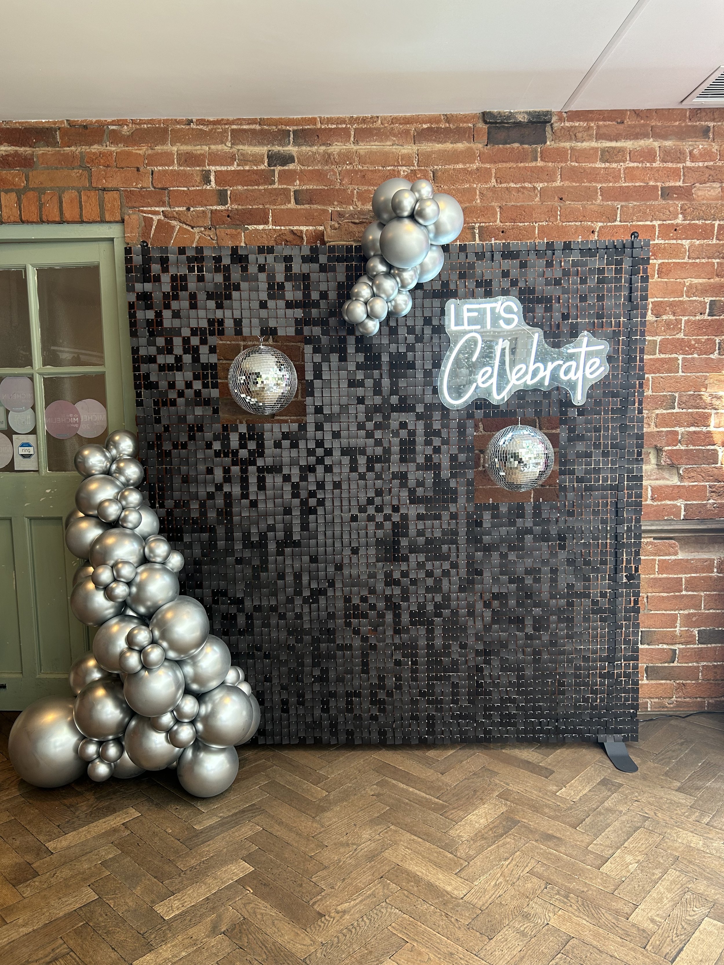 Decorative backdrop with black mosaic tiles, a cluster of silver balloons, two disco balls, and a neon sign that says 'Let's Celebrate' against a brick wall.