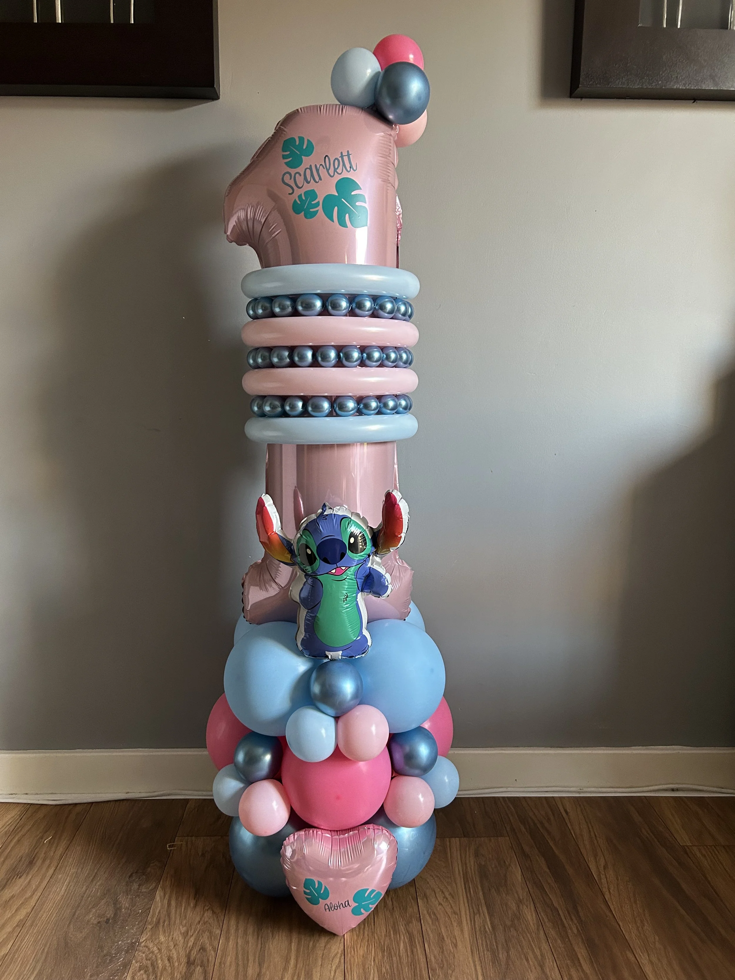 Colorful balloon arrangement with a pink '1' shaped balloon, other pink, light blue, and metallic gray balloons, a 'Lilo & Stitch' character balloon, and a pink heart-shaped balloon with the name 'Aloha' and a leaf graphic, set against a plain wall and hardwood floor.