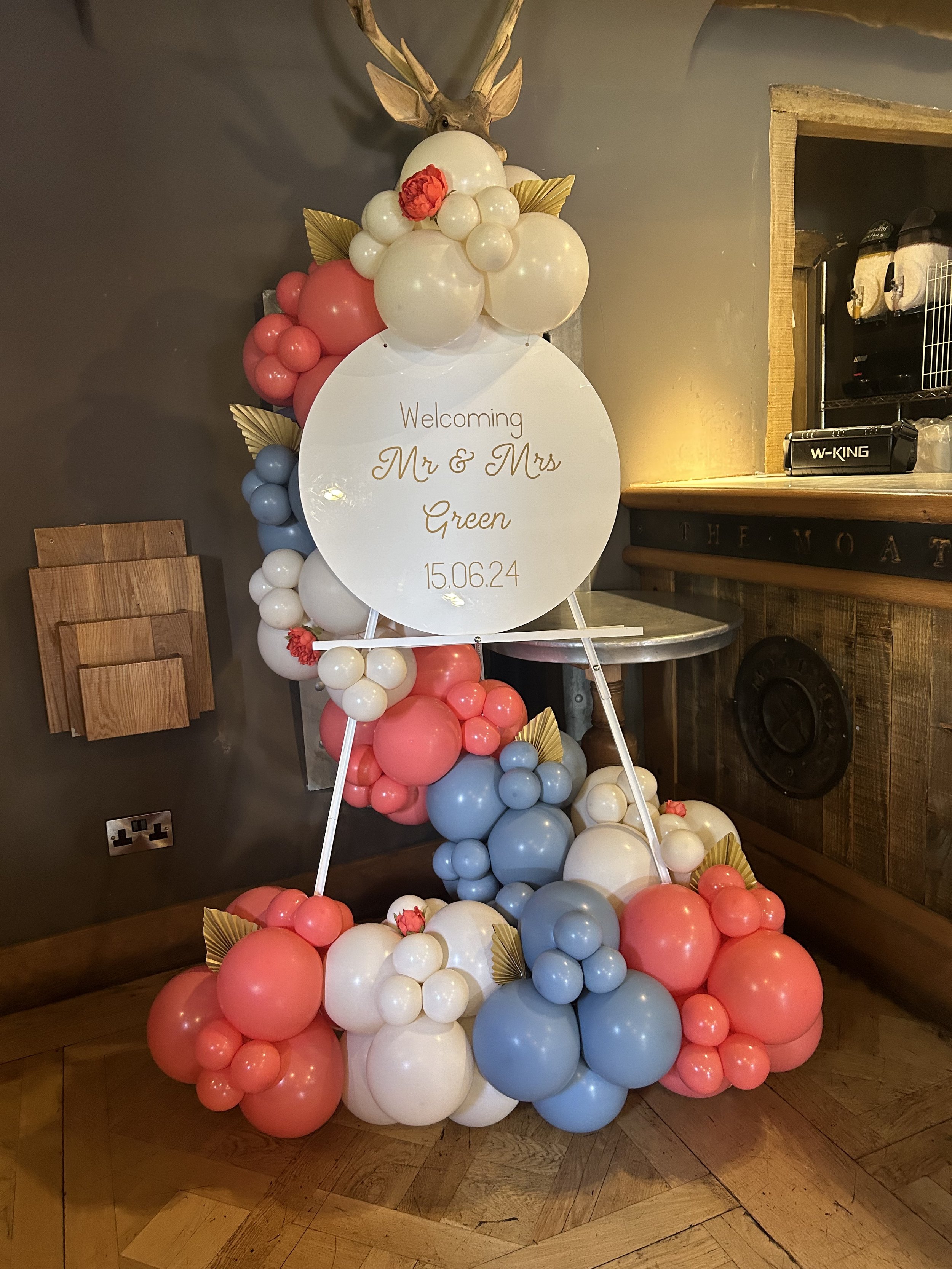 Balloon arrangement with pink, white, and blue balloons, adorned with gold leaves and a decorative reindeer head at the top, and a round sign welcoming Mr. & Mrs. Green, dated 15.06.24.