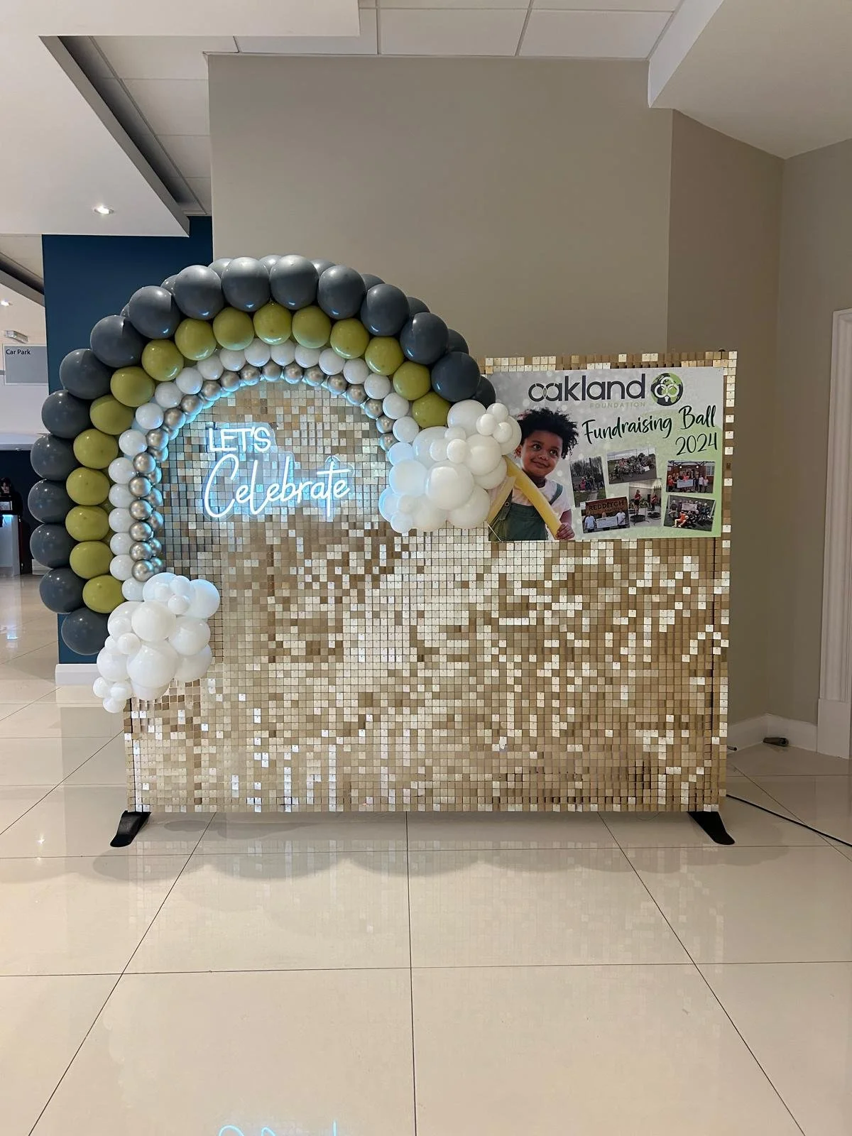 Decorative backdrop with balloons and a sign for Oakland Foundation fundraising event, 2024, with a neon sign that reads 'Let's Celebrate' and a poster featuring a young girl.