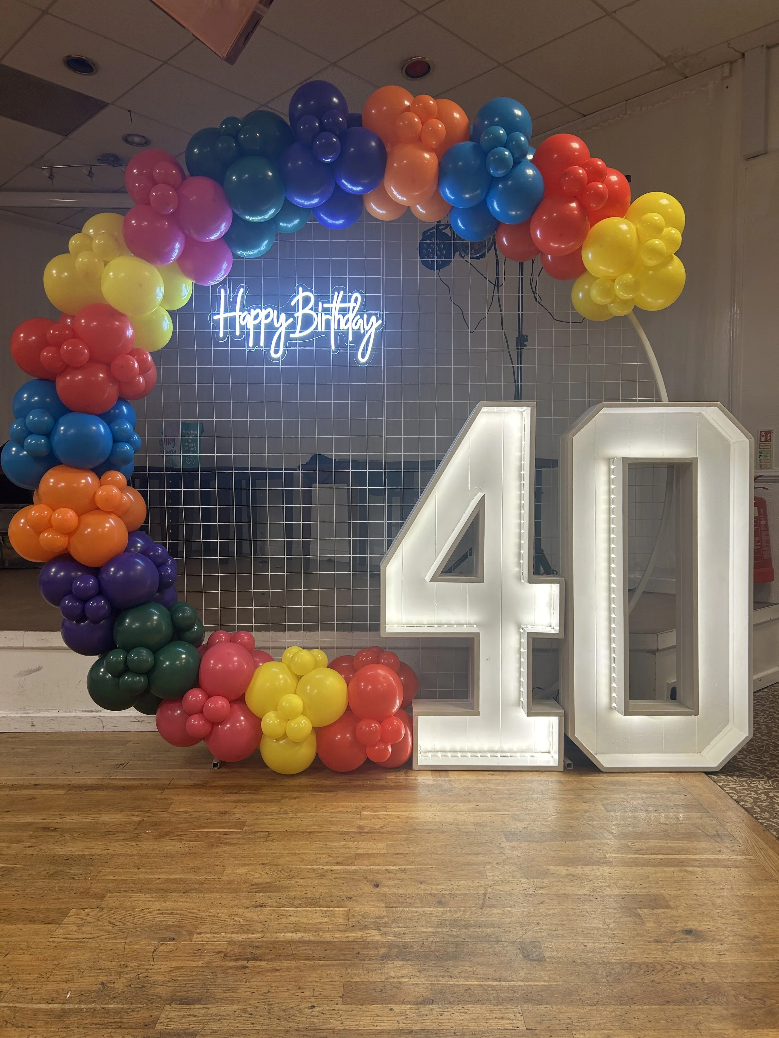 A birthday celebration setup with a balloon arch in various colors, a white illuminated number 40, a grid backdrop with a neon "Happy Birthday" sign, and a wooden floor.