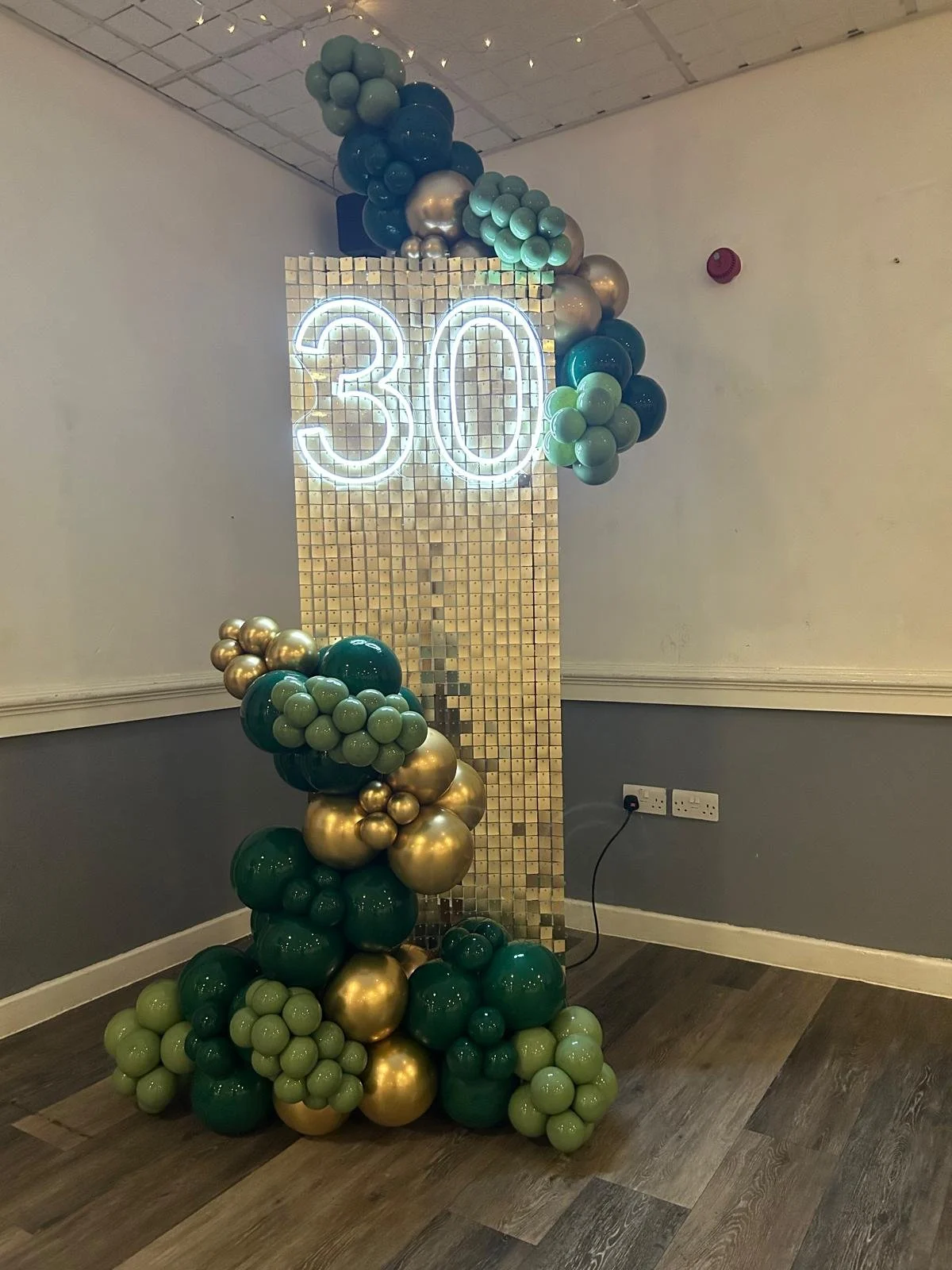 A decorative balloon display with clusters of green, gold, and silver balloons arranged around a illuminated '30' sign on a mirrored mosaic background.