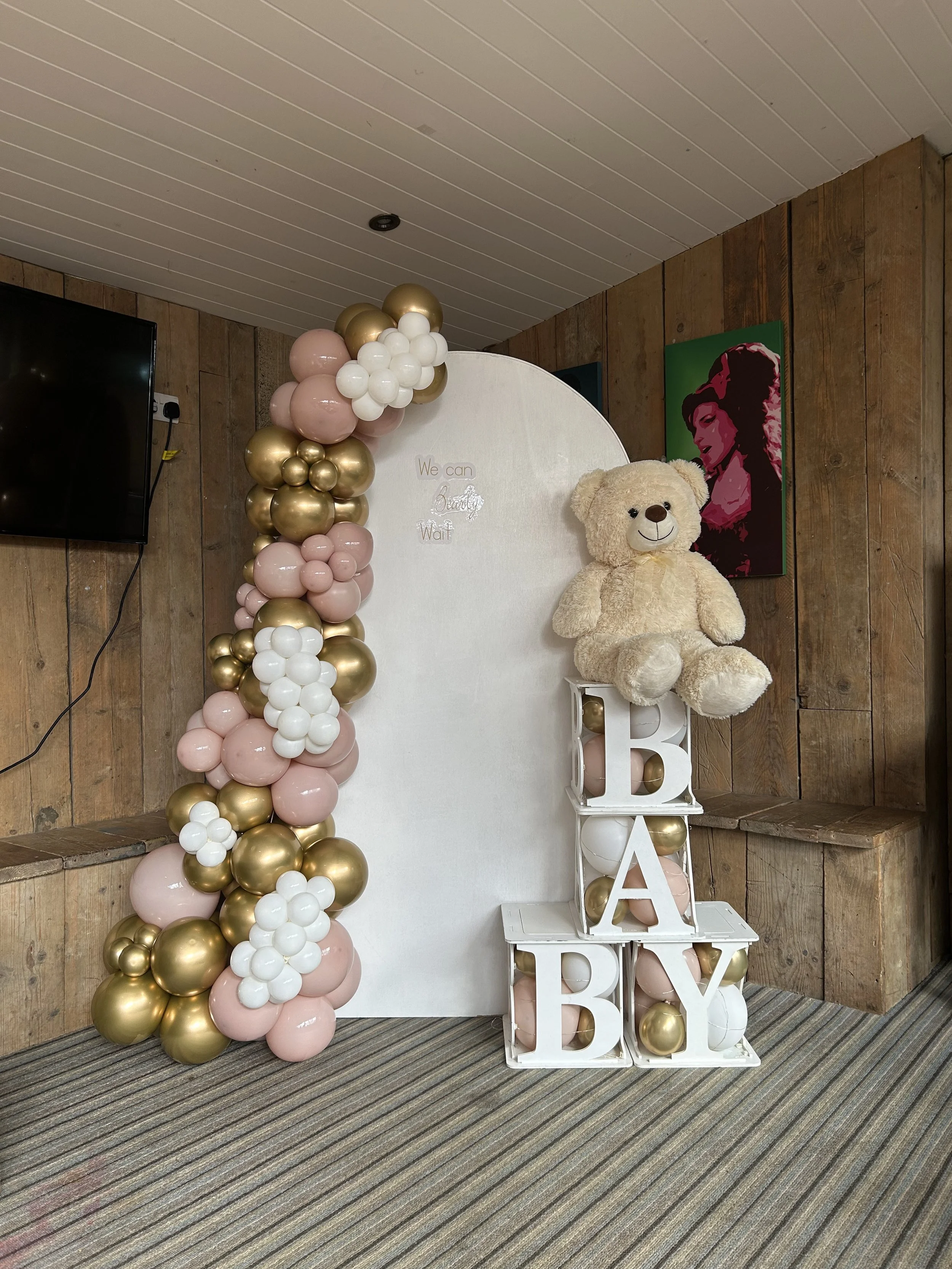 Decorative setup with pink, white, and gold balloons arranged around a white backdrop that reads 'We can be beautiful'. Beside the backdrop, there is a large teddy bear sitting on a white stand with large white letters 'B', 'A', and 'B' in front of the stand. A colorful portrait of a woman is on the wooden wall behind the setup.