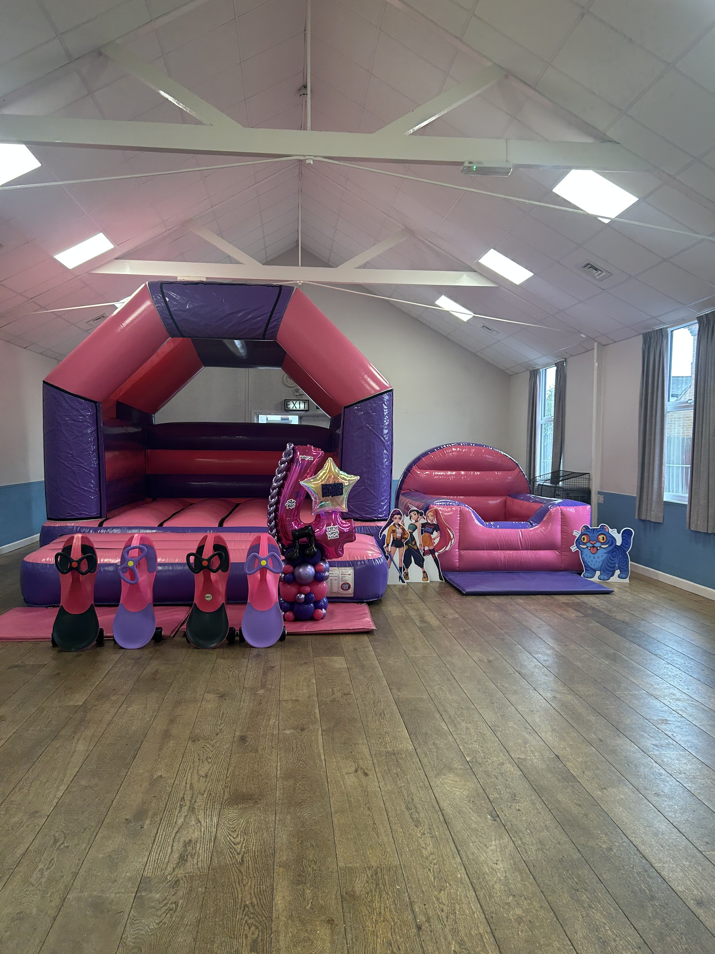Indoor play area with pink and purple inflatable bounce house, a pink inflatable slide, and party decorations including balloons and cardboard cutouts of cartoon characters. The room has hardwood floors, high ceiling with white beams, and large windows with curtains.