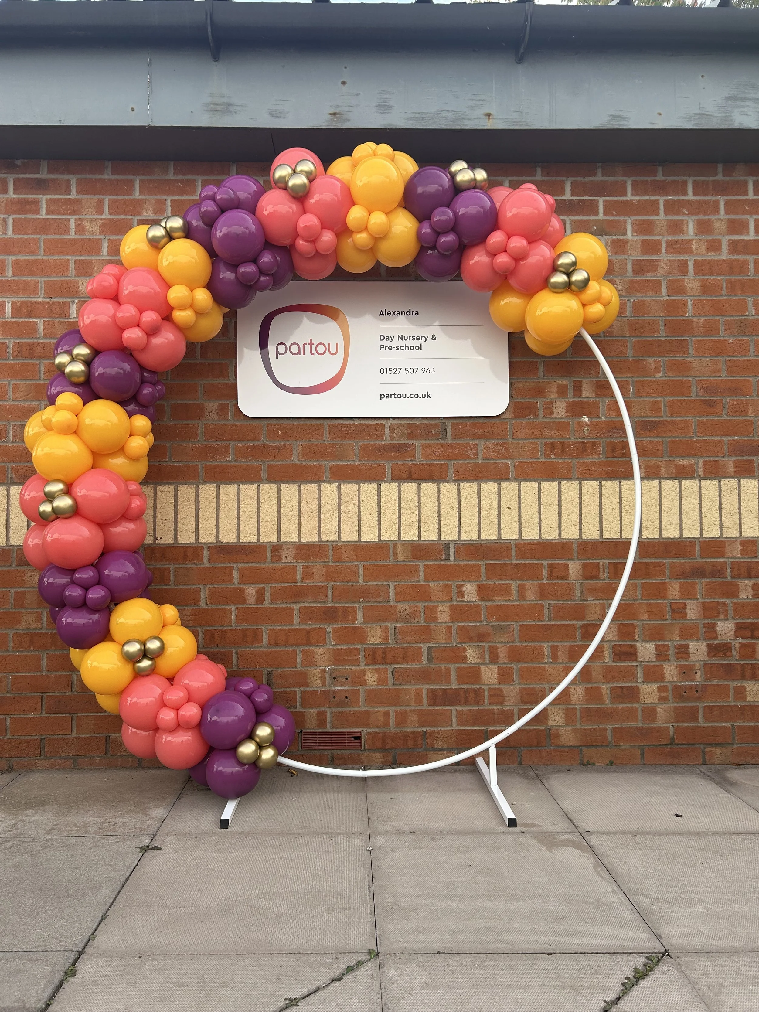Balloon arch with pink, purple, yellow, and gold balloons forming an arc, positioned in front of a brick wall and a white sign for Partou Day Nursery & Pre-school, with a white stand and cord on the ground.