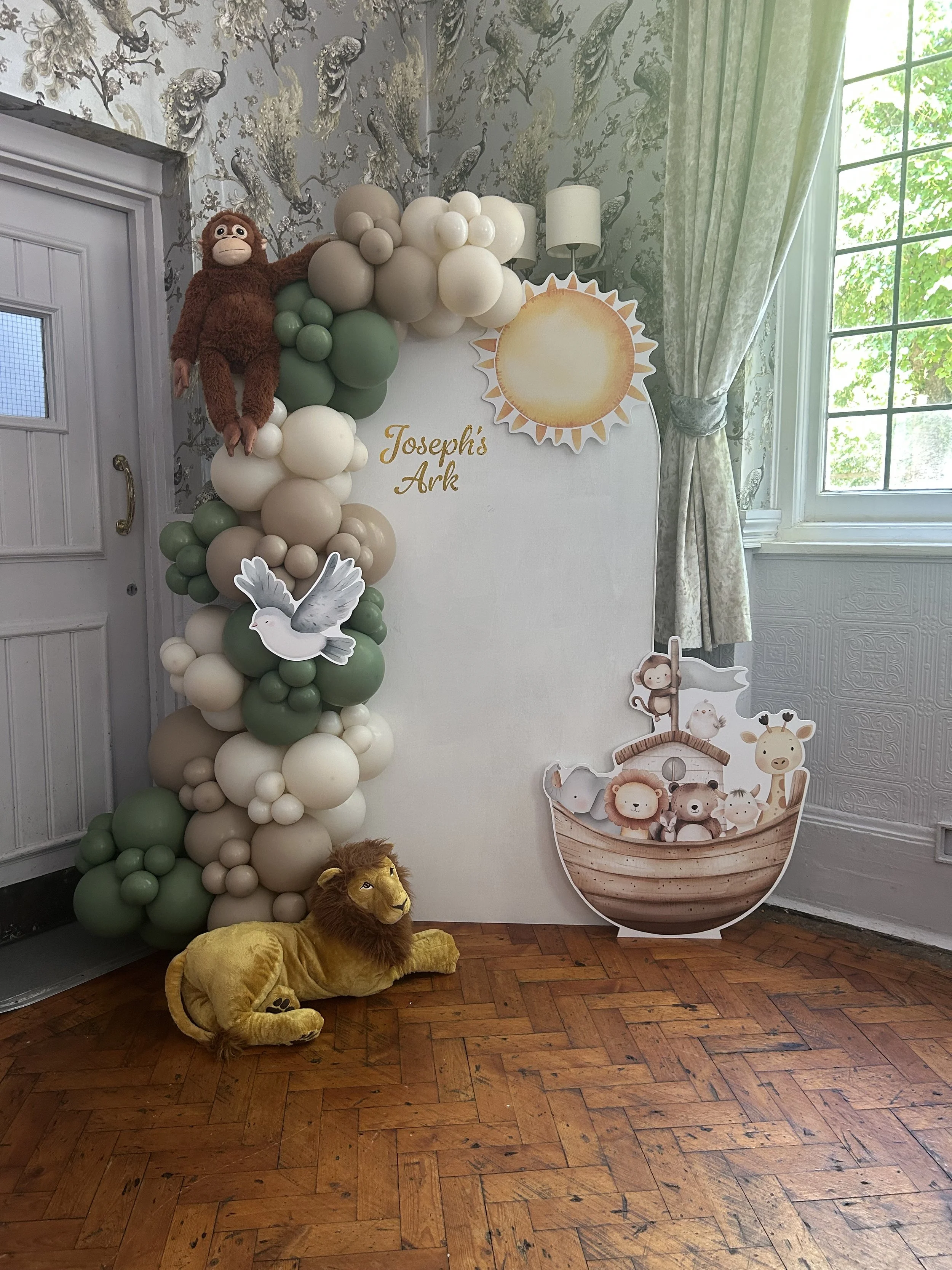 Decorative setup with a plush lion on the floor, a backdrop with balloons and cutouts of a monkey, birds, and safari animals, including a giraffe, in a room with wooden flooring, patterned wallpaper, and a window with curtains.