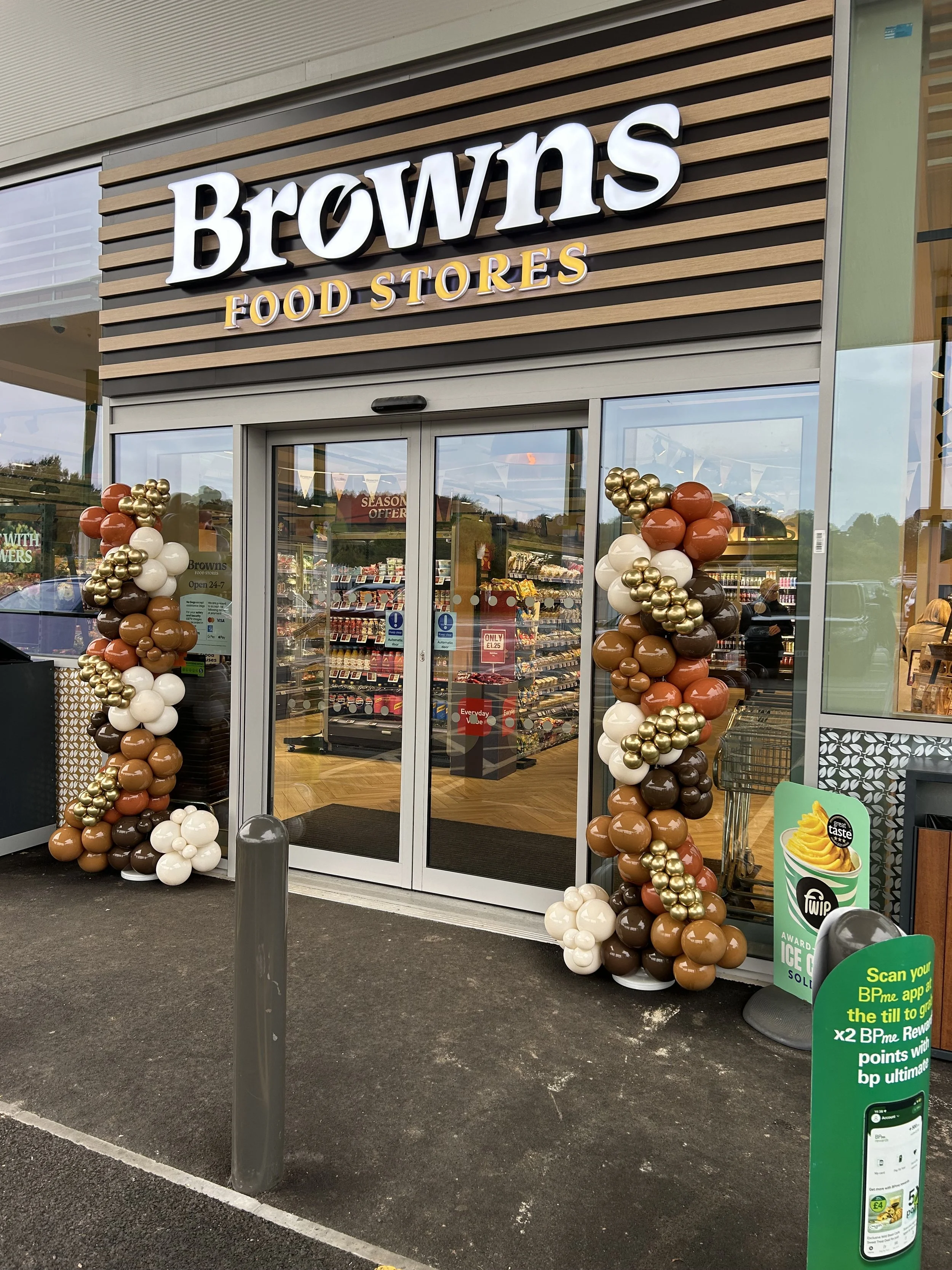 Exterior view of a Browns Food Stores entrance decorated with balloon garlands in brown, white, gold, and orange. Store sign is illuminated above the entrance, with glass doors showing store interior and shelves filled with products.