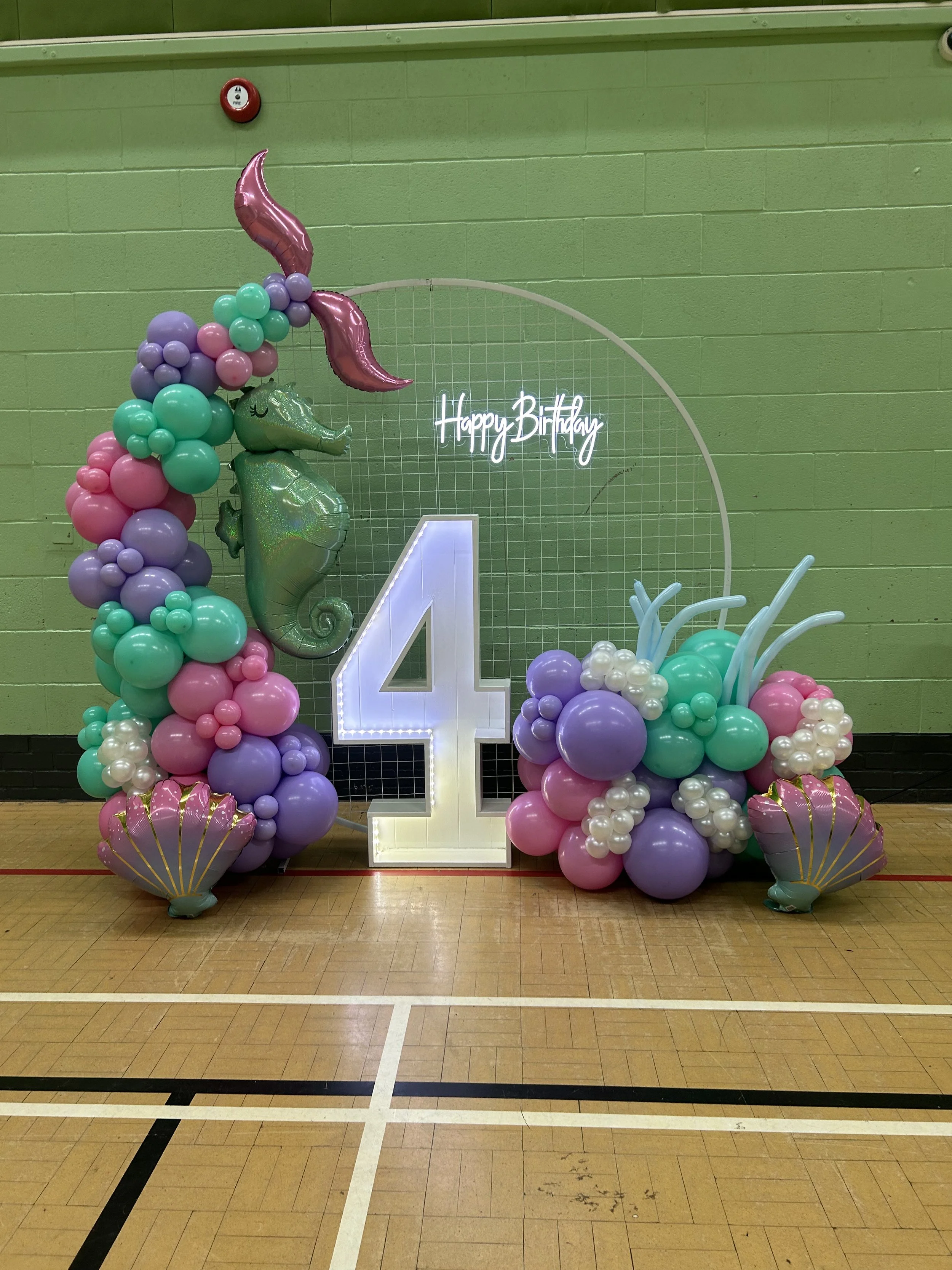 Decorative birthday party display featuring pastel-colored balloons arranged around a large illuminated number 4, with a neon sign that says 'Happy Birthday', set against a green wall in a gymnasium or indoor sports facility.
