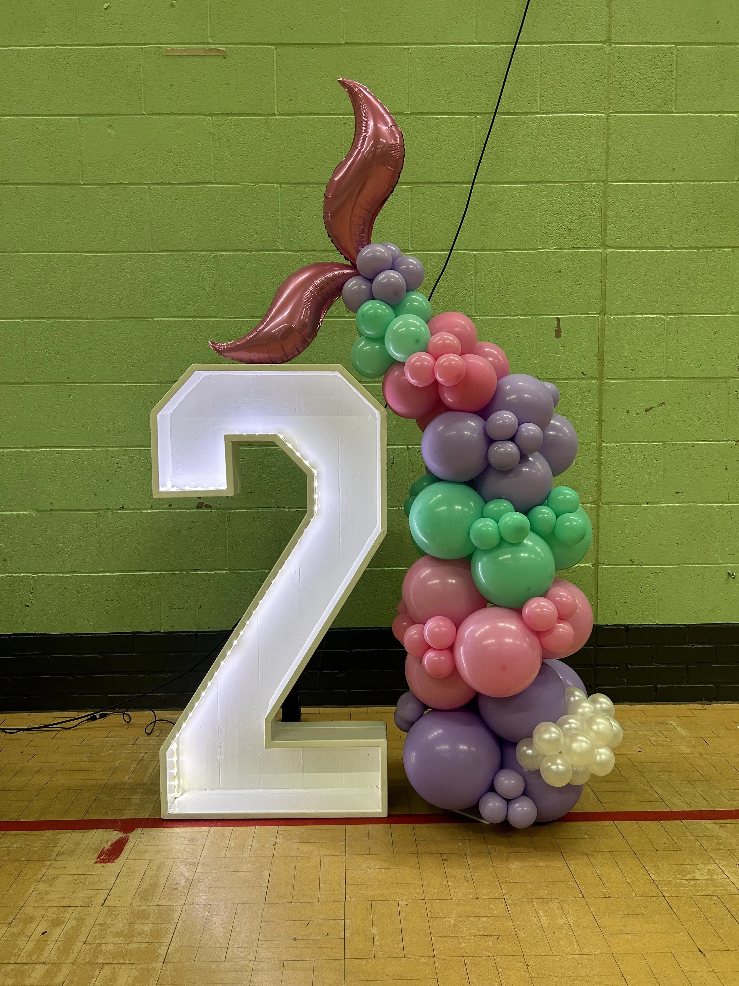 Decorative birthday display featuring a large illuminated number 2, pink, purple, green, and white balloons arranged in a cascading design, set against a lime green brick wall in an indoor gymnasium.