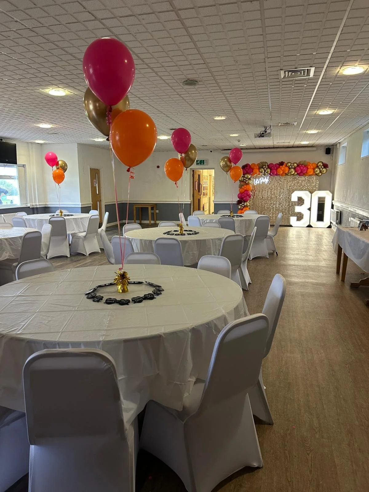 A decorated event hall with round tables covered in white tablecloths, topped with balloon centerpieces in pink, orange, and gold. The back wall features a balloon arch in similar colors, large illuminated number '30', and a golden sparkly backdrop.