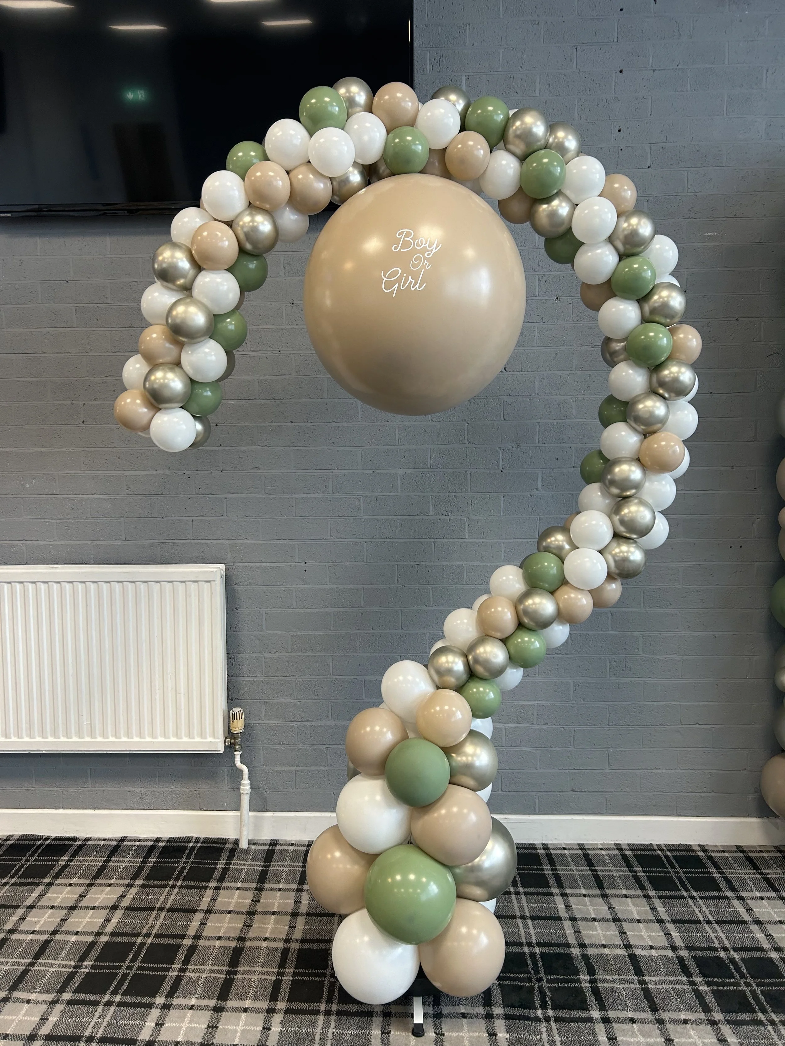 Decorative arch made of balloons in shades of white, cream, green, and metallic gold, with a large cream-colored balloon in the center that says 'Boy Or Girl' in white script, set against a gray brick wall.