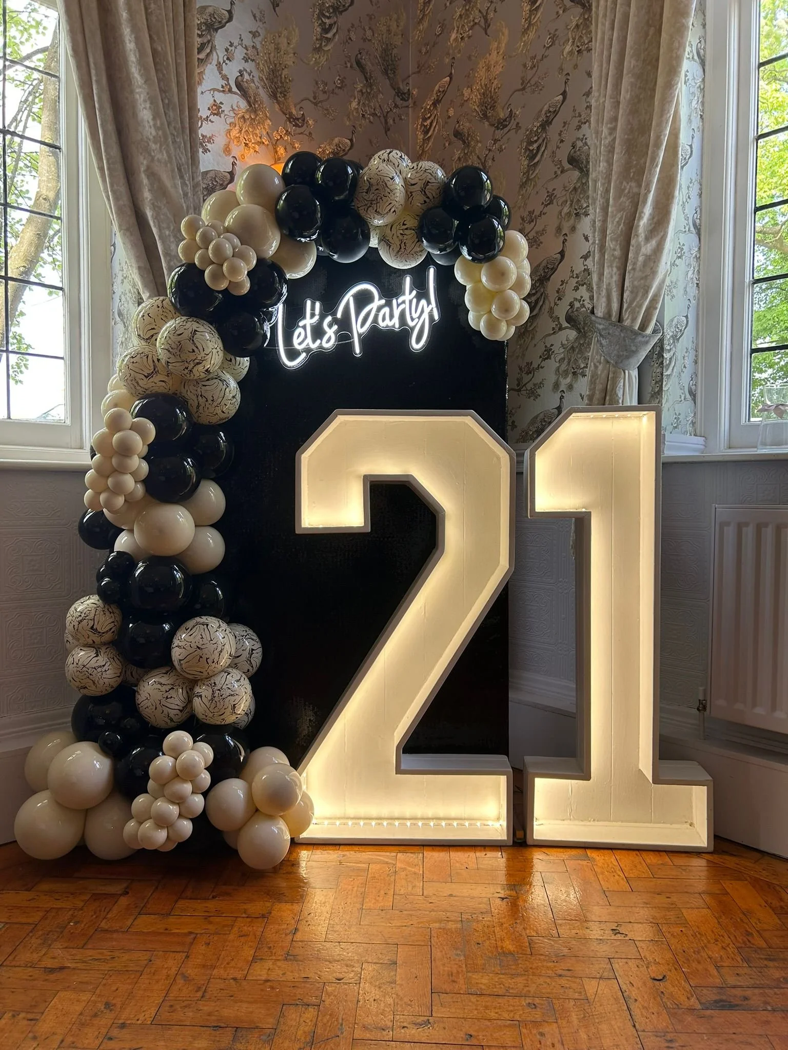 Decorative photo backdrop with a balloon arch on the left and illuminated large number '21' on the right, against a floral wallpaper with peacock motifs, in a room with windows and wooden floor.
