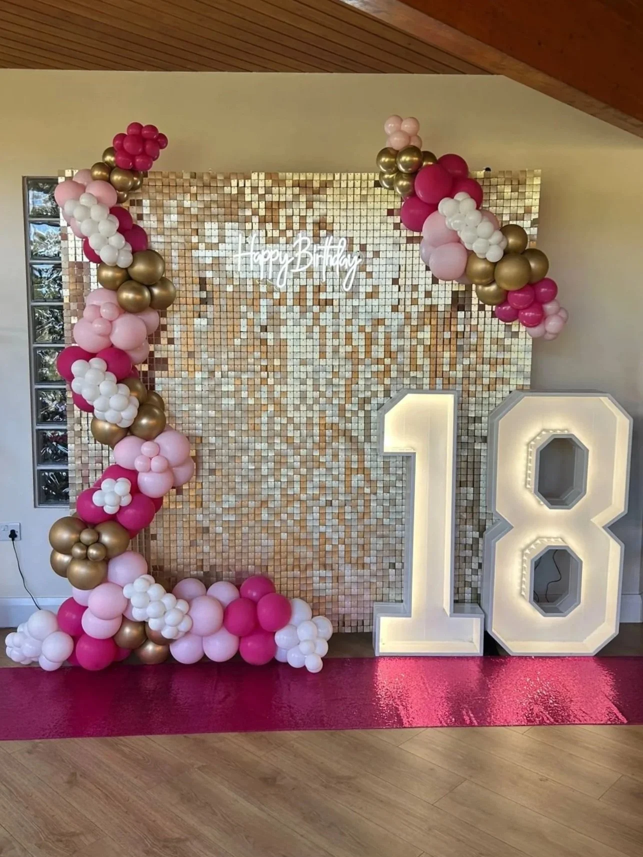 Decorative birthday backdrop with balloon arch, large illuminated number 18, and a sparkly gold tiled wall. The backdrop features a neon sign that says 'Happy Birthday' with gold and pink balloons surrounding it.