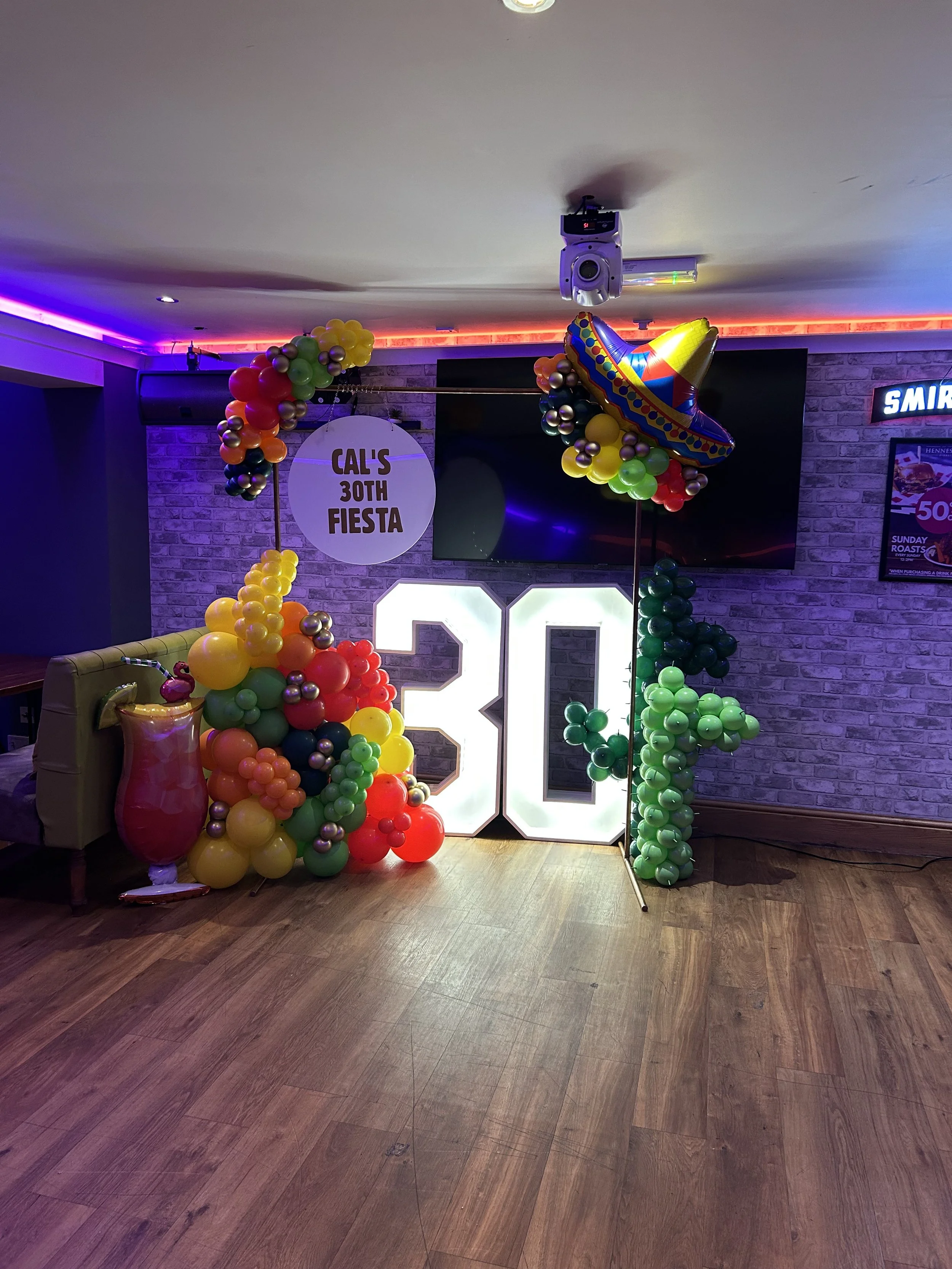 Decorations for a birthday party with colorful balloons, a large illuminated number 30, a sign reading 'Cal's 30th Fiesta,' and a Mexican sombrero balloon.