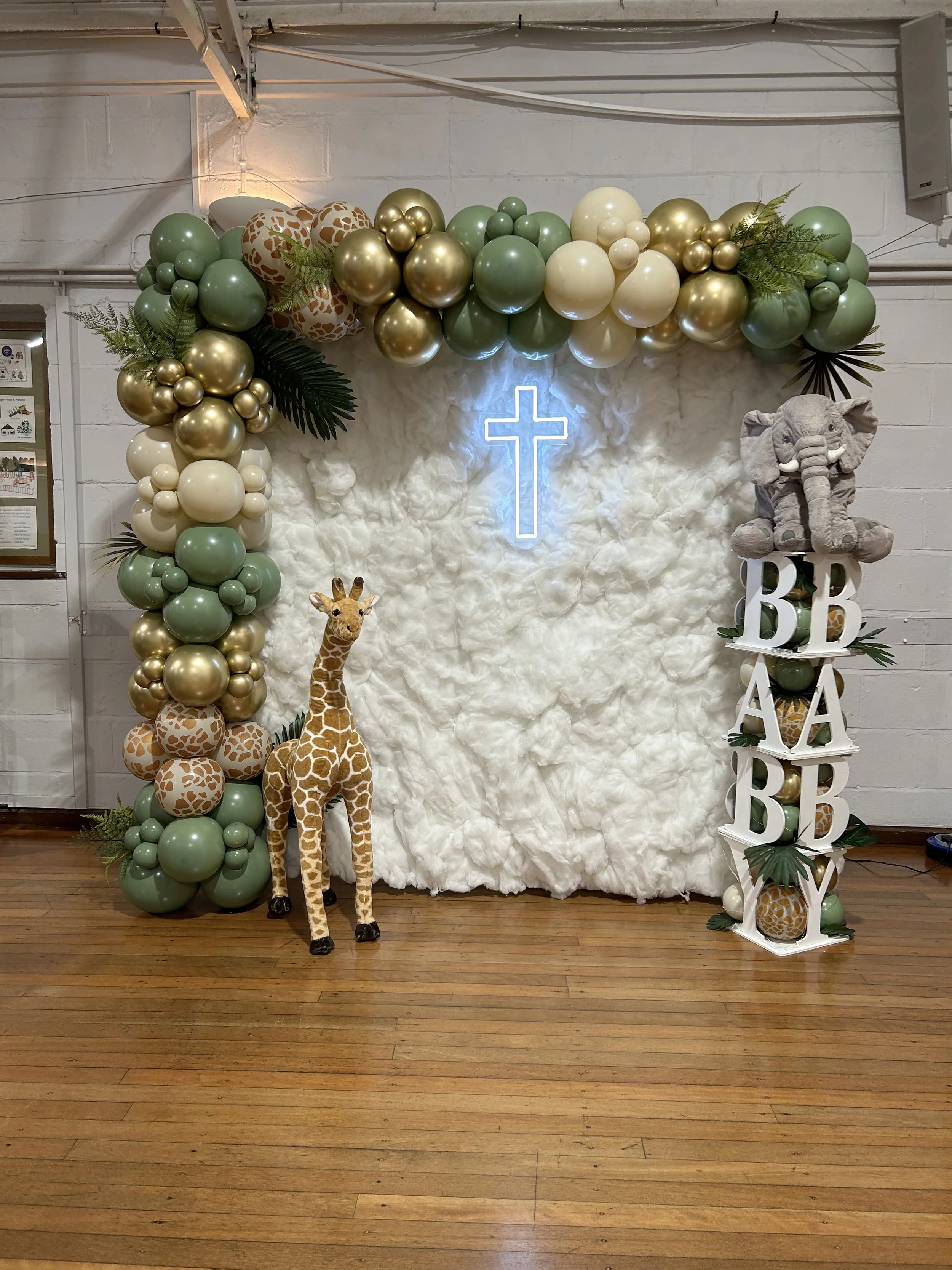 Decorative setup with a white fluffy background, a cross neon sign, and arrangements of green, gold, cream, and balloon giraffe-themed decorations. Includes plush giraffe, greenery, and the words 'BABY',