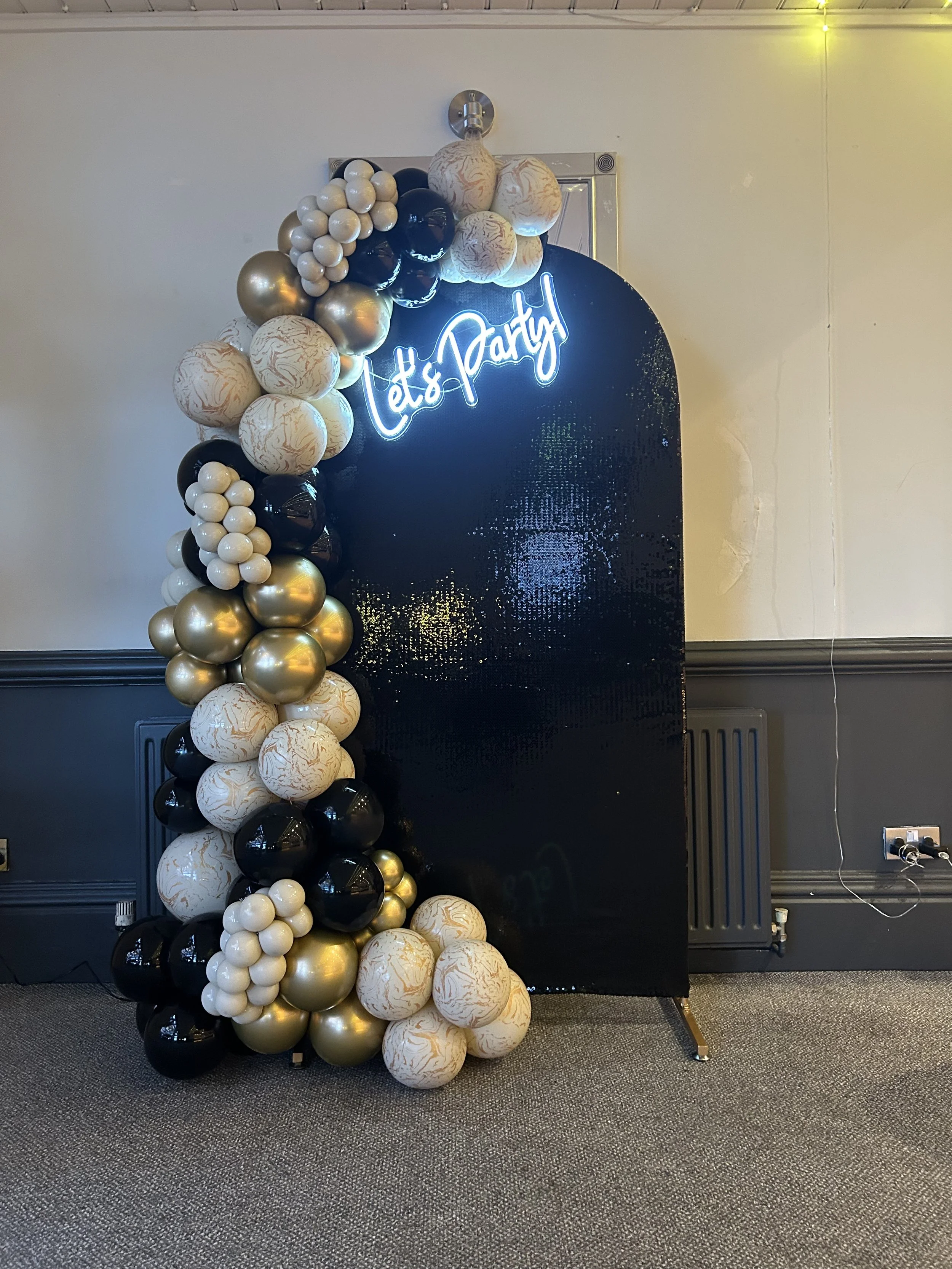 Decorative balloon arch with black, white, gold, and marbled balloons, set against a black sequin backdrop with a neon sign that reads "Let's Party" in cursive.