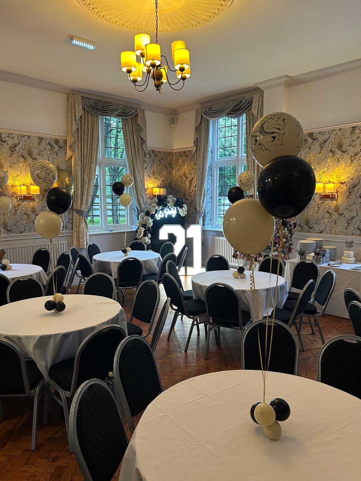 Decorated event room with black and white balloons, round tables with white tablecloths, and a large illuminated number 21 in front of windows with beige curtains.