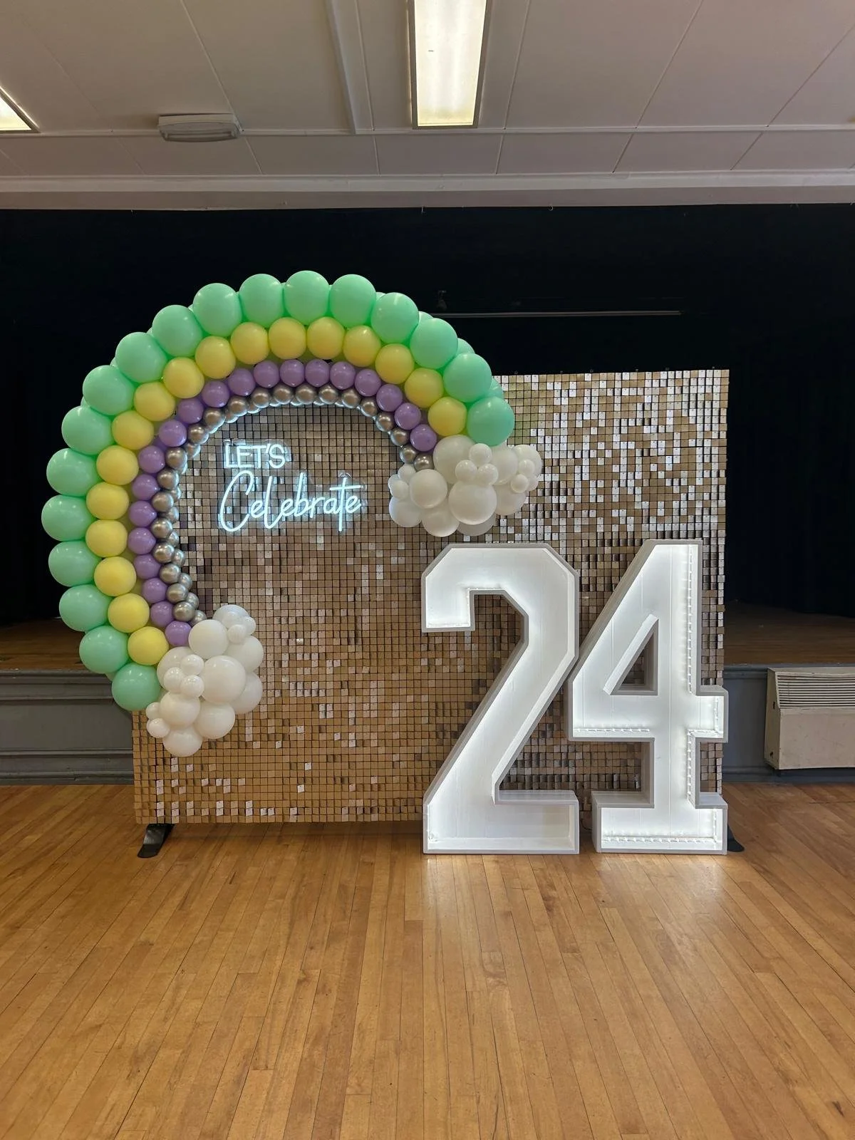 Decorative birthday celebration backdrop featuring a balloon rainbow with pastel colors, a neon sign that says 'Let’s Celebrate', large illuminated number 24, and a mosaic tiled background on a wooden floor.
