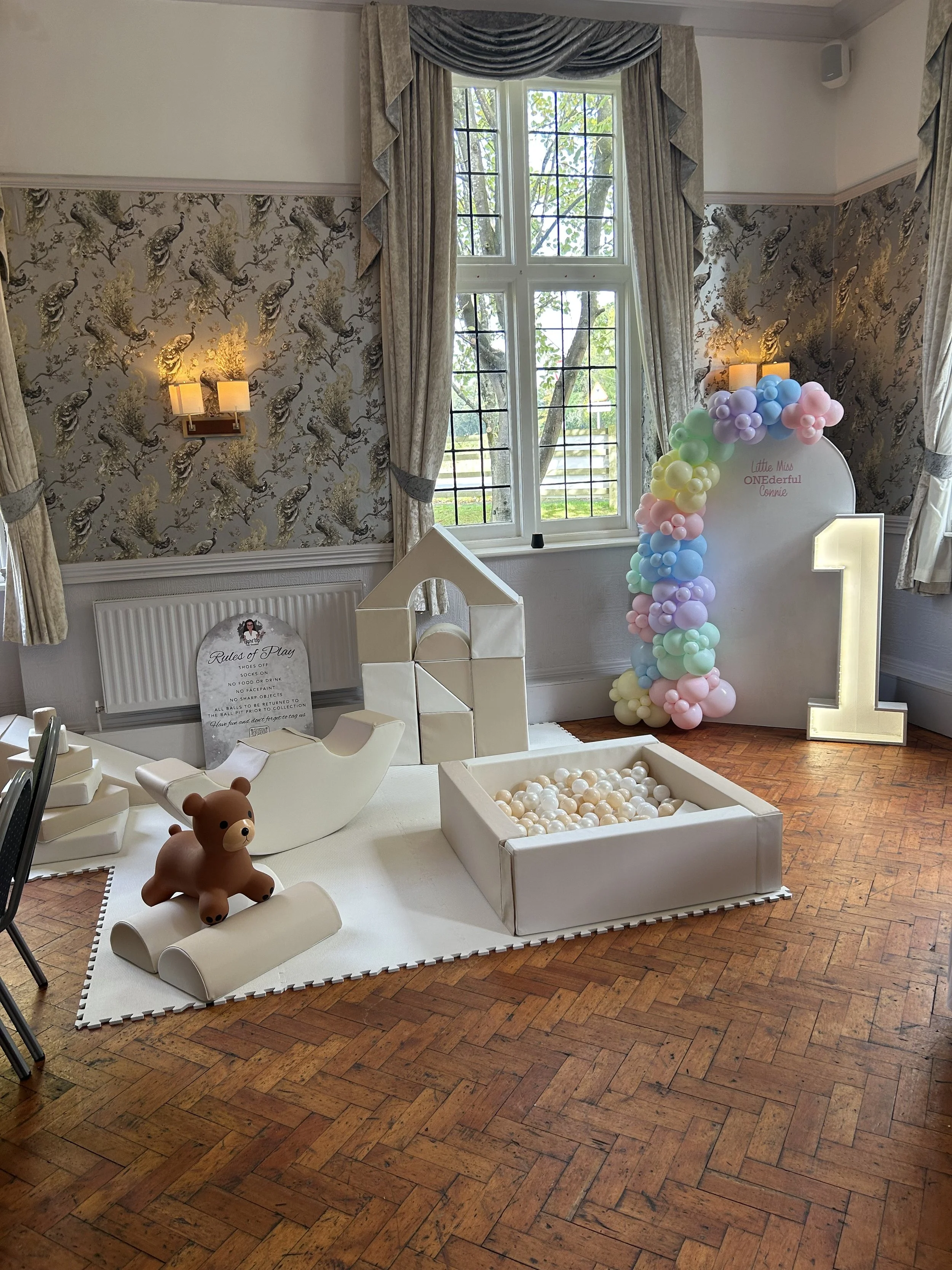 A children's play area with a teddy bear, ball pit filled with white balls, and decorative structures on a white mat in front of a window with curtains. A large illuminated number one and a balloon arch with pastel-colored balloons are also present.