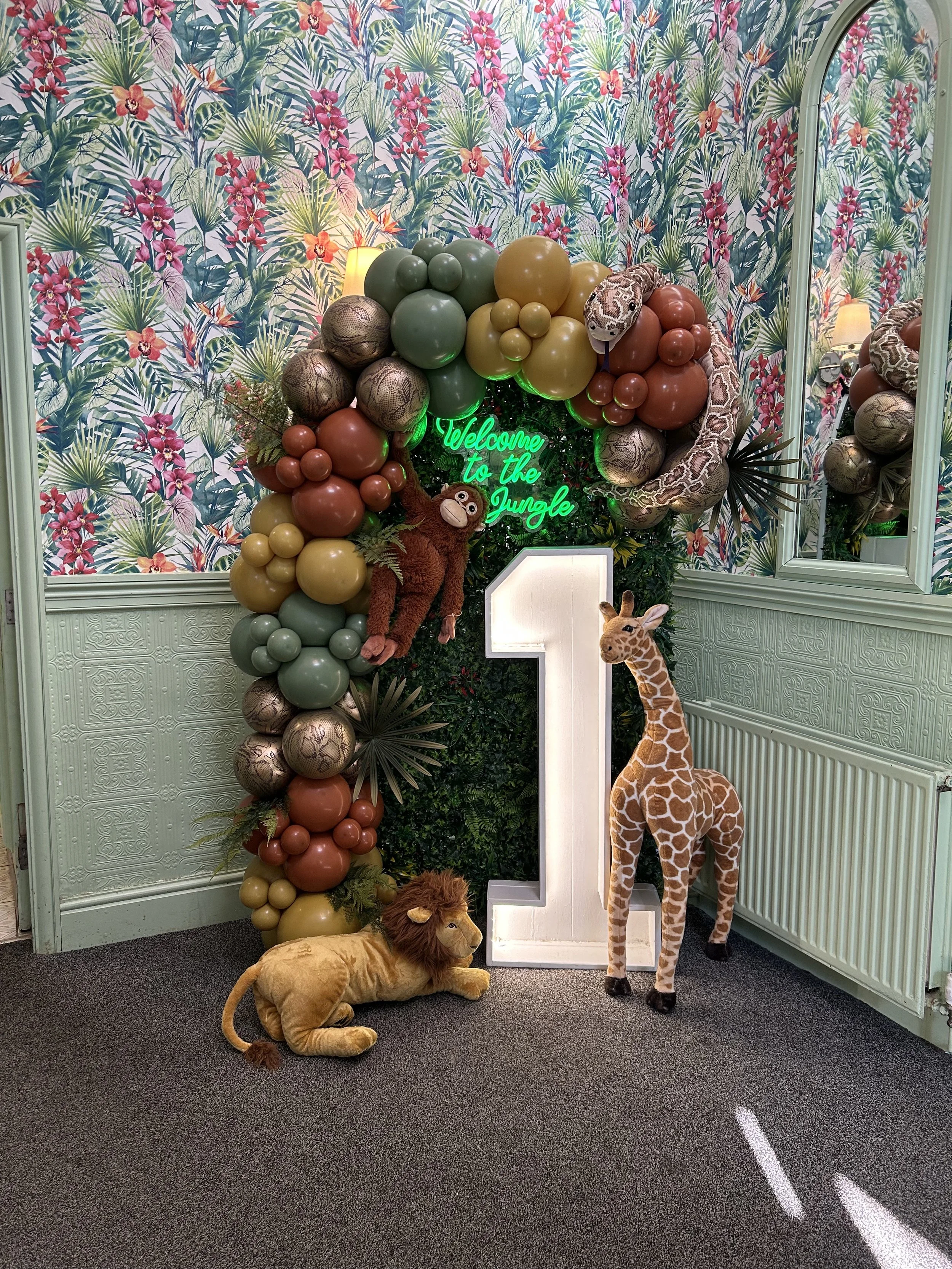 Decorative jungle-themed backdrop with balloons, plush animals, and a neon sign that reads "Welcome to the Jungle." Plush lions, giraffe, and snake figures are placed in front of the backdrop, along with a large illuminated white number one.