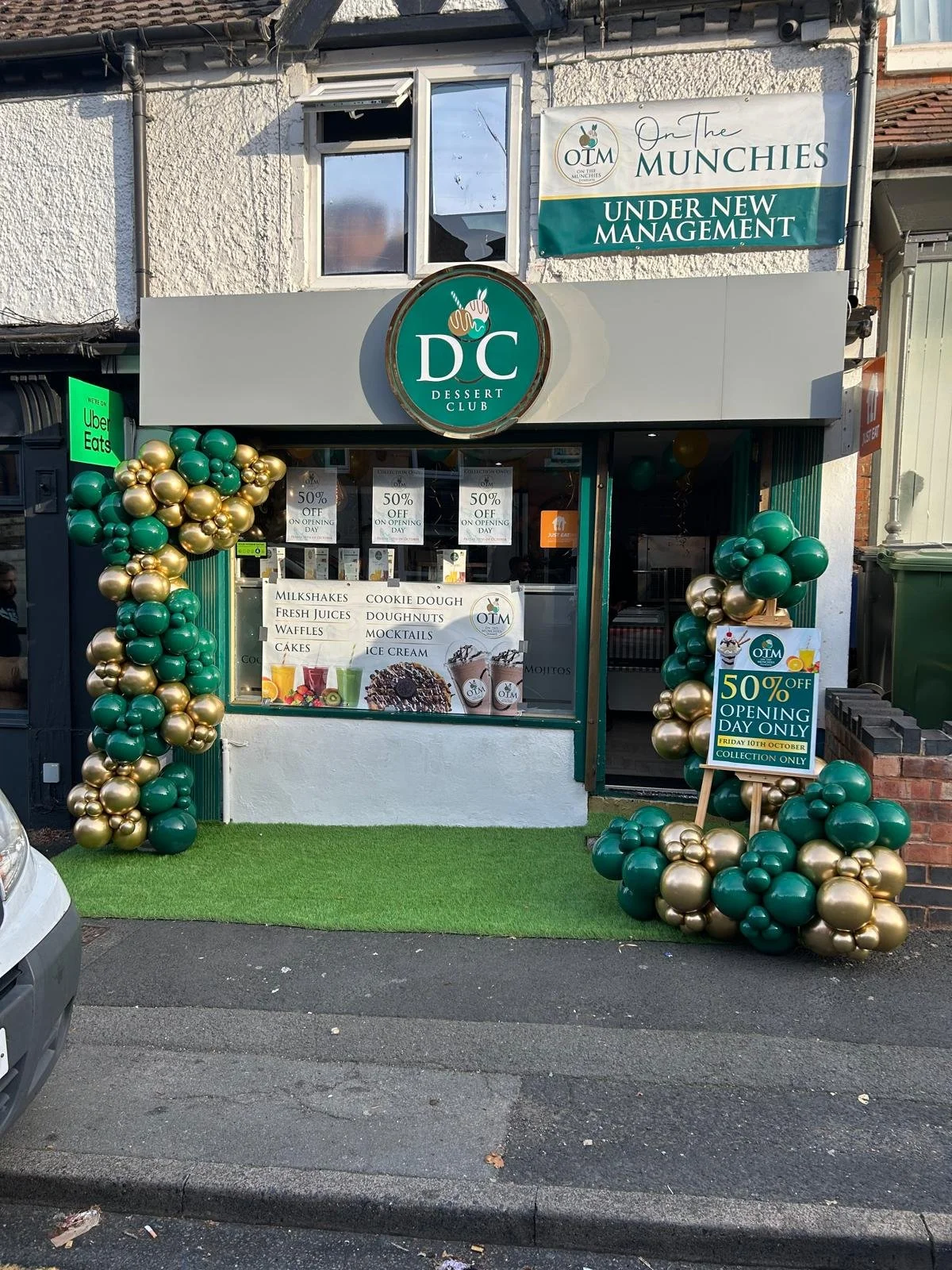 storefront decorated with green and gold balloons, advertising a 50% discount for opening day of a dessert shop called DC Dessert Club, featuring menu items like milkshakes, cookie dough, fresh juices, doughnuts, waffles, mocktails, cakes, and ice cream.