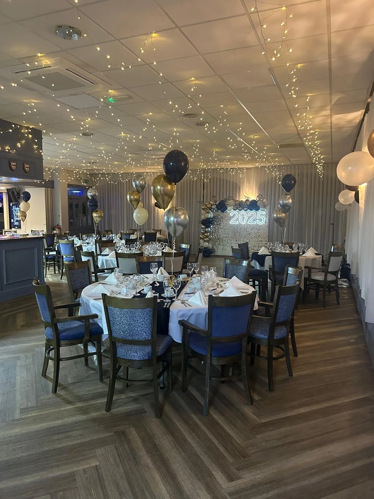 A decorated banquet hall with round tables, balloons, fairy lights, and a backdrop displaying '2025' for a New Year celebration.