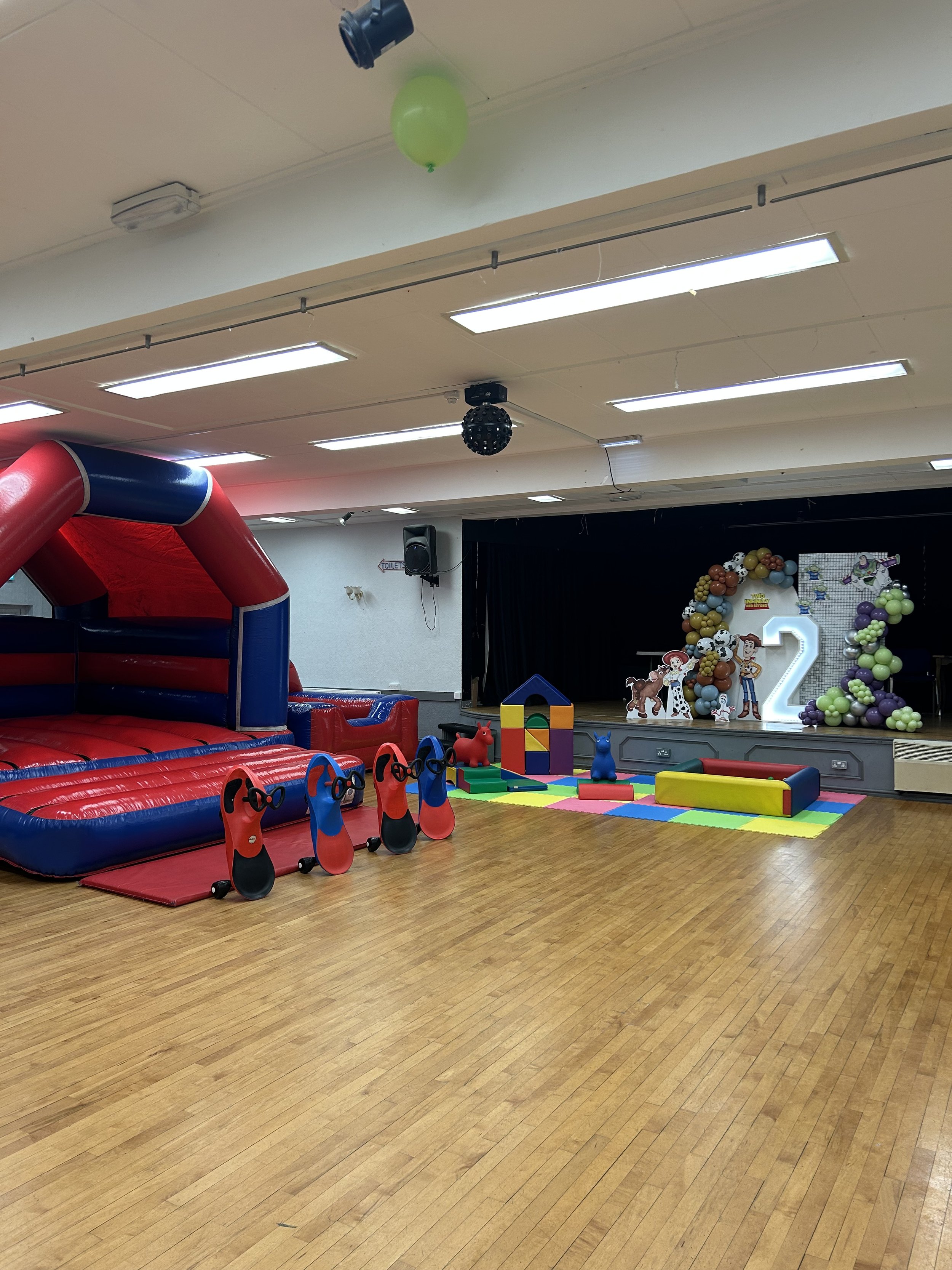 An indoor children's birthday party setup with a bounce house, colorful toys, and a stage decorated with balloons and cartoon characters.