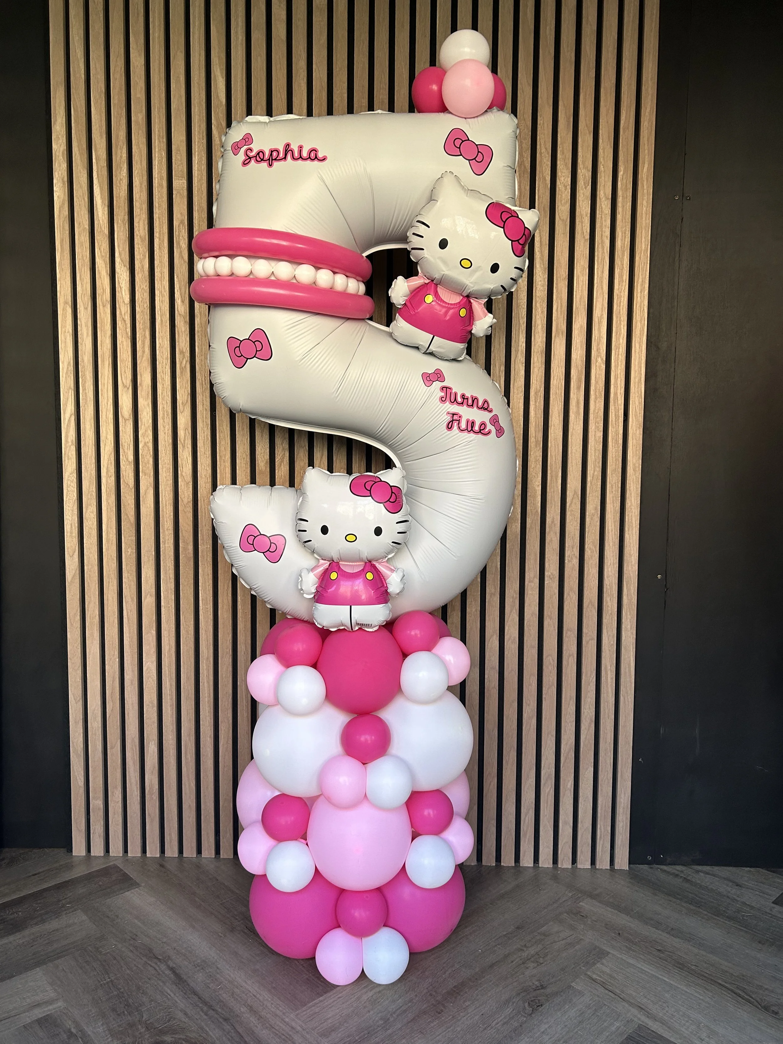 Pink and white balloon display shaped like a number 3 with Hello Kitty decorations, including a large bow and a smaller plush Hello Kitty figure, on a wooden-paneled wall.
