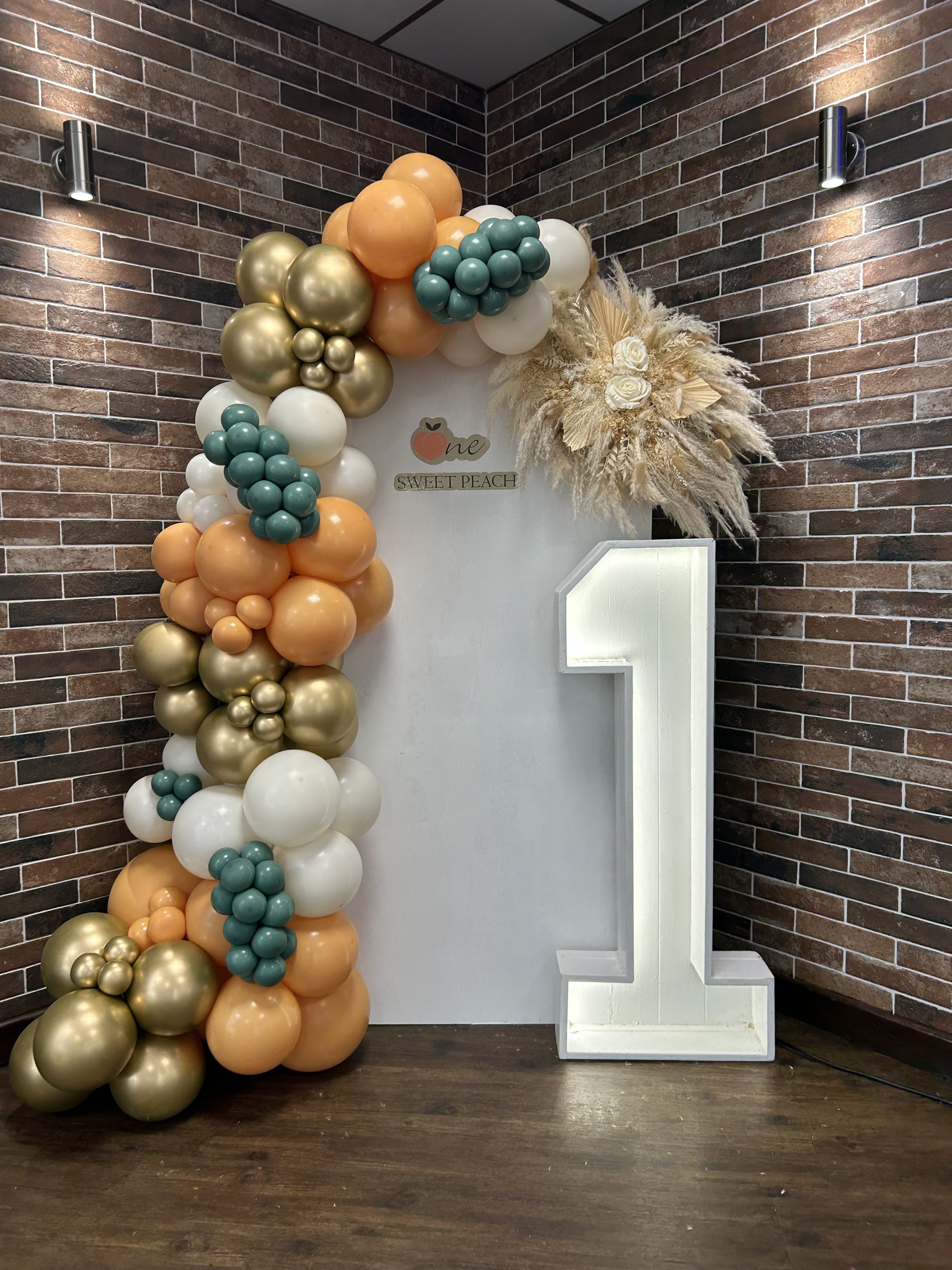 Celebration backdrop with a balloon arch and a white letter 'E' with a floral arrangement. The balloons are in shades of gold, peach, white, and teal, arranged in a cascade style on a brick wall background.