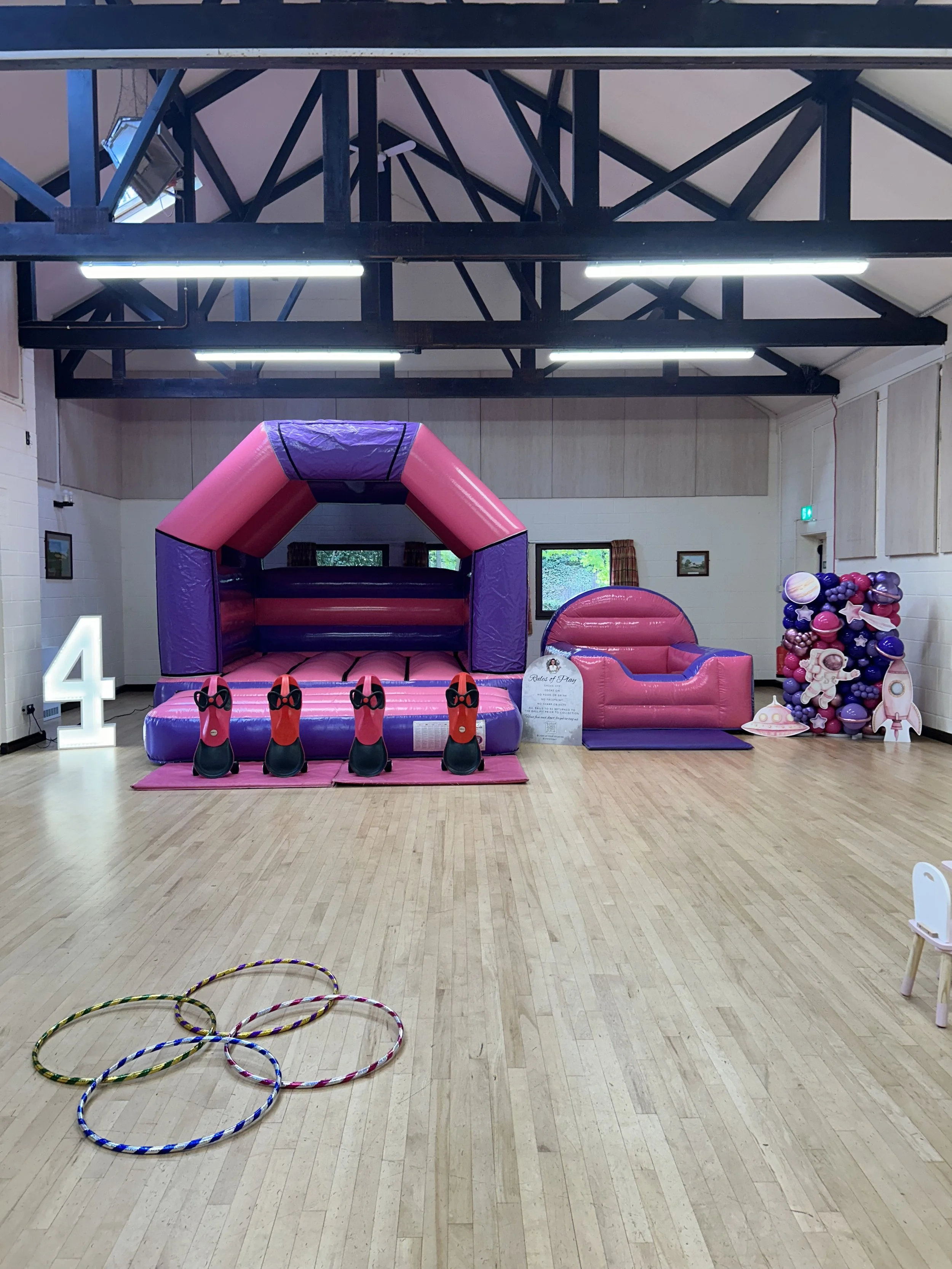 Indoor space with wooden flooring, a pink and purple inflatable obstacle course, a small pink slide, colorful hula hoops on the floor, and a balloon display on the right side. There is a large white illuminated number 4 on the left.