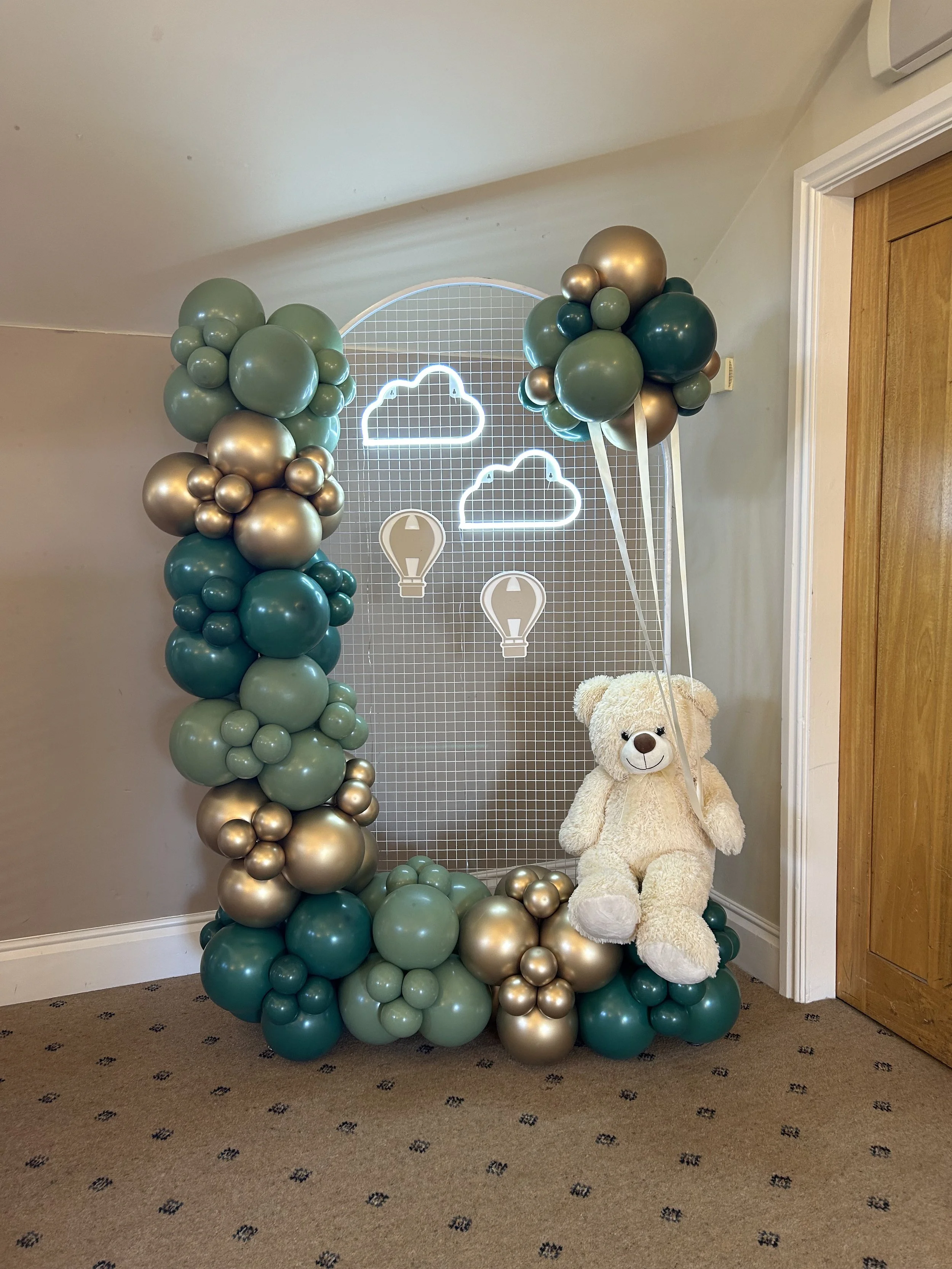 A decorative arrangement of green, gold, and teal balloons forming an arch and columns, with a large teddy bear sitting on the floor next to the balloons, in front of a grid backdrop with neon cloud and hot air balloon icons.