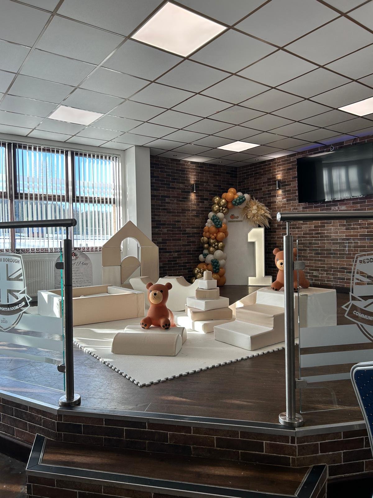 Indoor celebration setup for a first birthday with a white platform, teddy bear figures, a large white number one, and a balloon arch against a brick wall, near a window with vertical blinds.