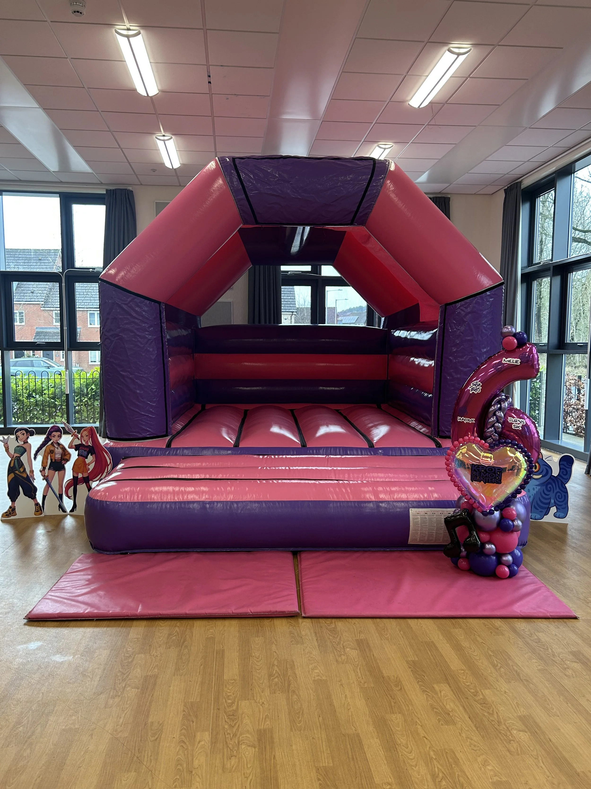 A large indoor bounce house with pink and purple walls, a pink padded entrance, and decorations featuring characters from the show 'KPOP Demon Hunters'. There are large windows with views of houses and trees outside.