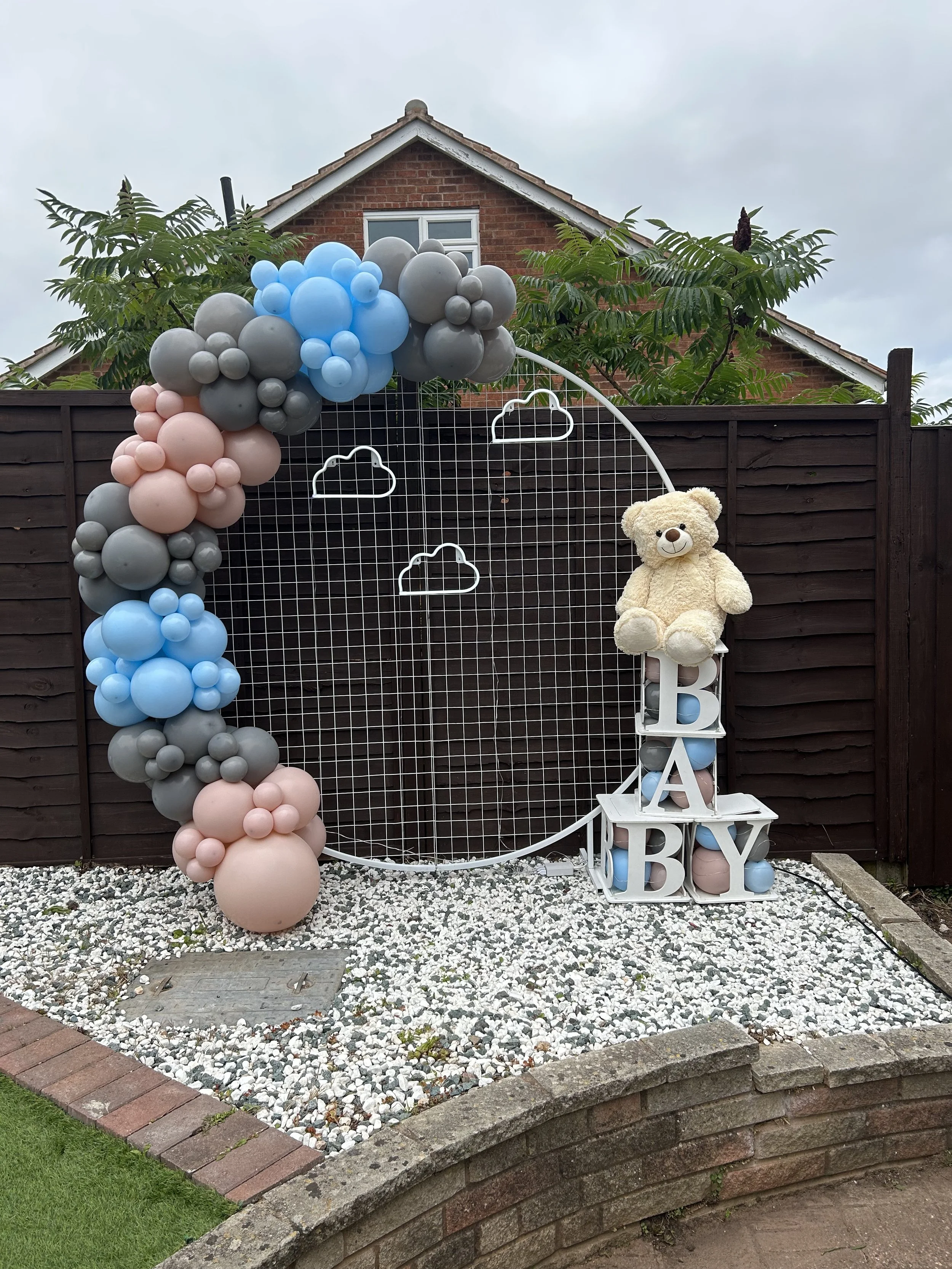 Outdoor decoration with pastel-colored balloons forming an arch around a white metal frame, a plush teddy bear on a white stand, and large decorative blocks spelling 'BABY', set against a dark wooden fence and a brick house with trees in the background.