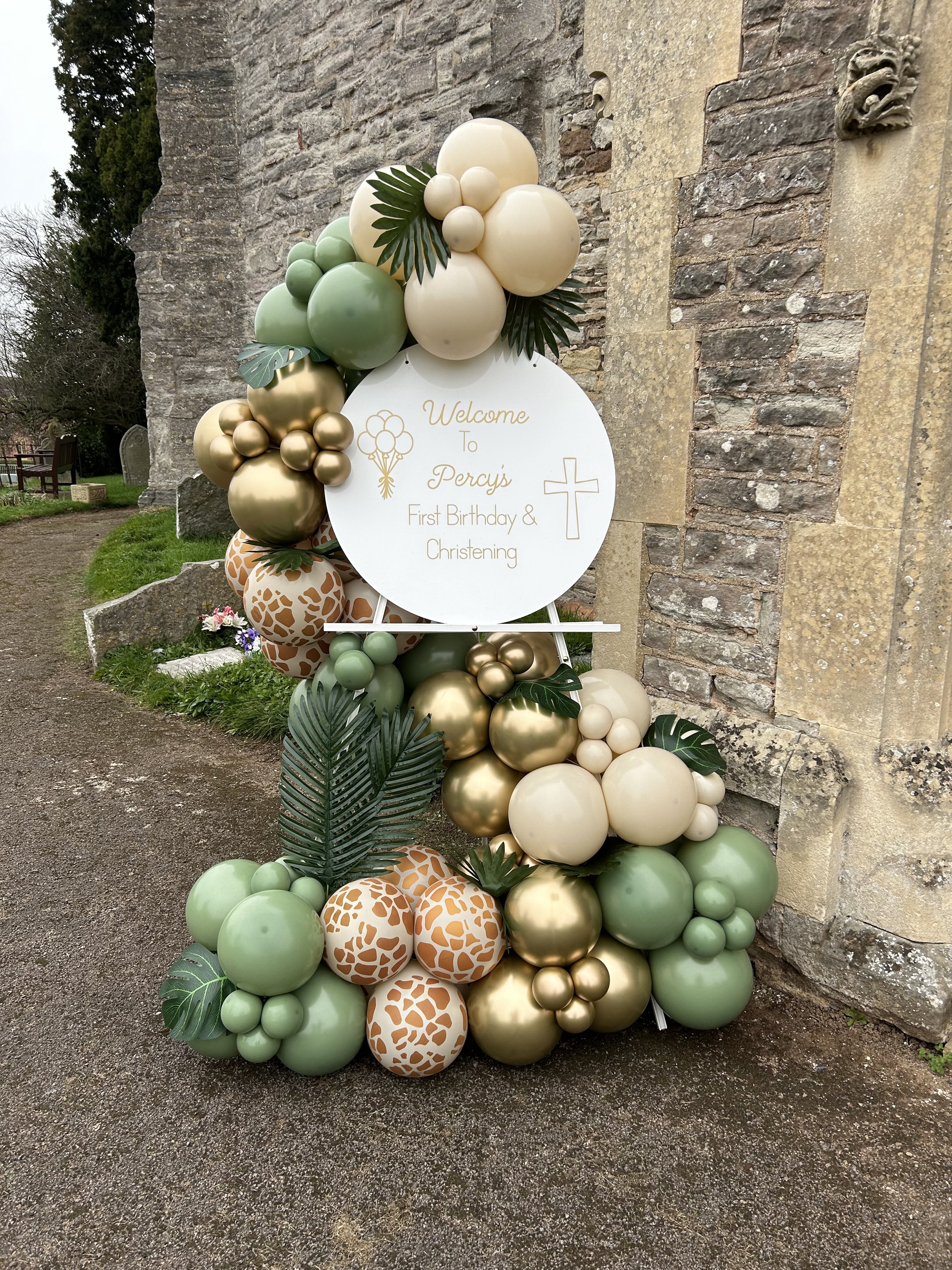 Decorative cluster of balloons in green, gold, cream, and giraffe print patterns arranged in front of a stone wall, with a white circular sign that reads, "Welcome To Percy’s First Birthday & Christening" adorned with balloons, balloons, balloons, and tropical leaves.
