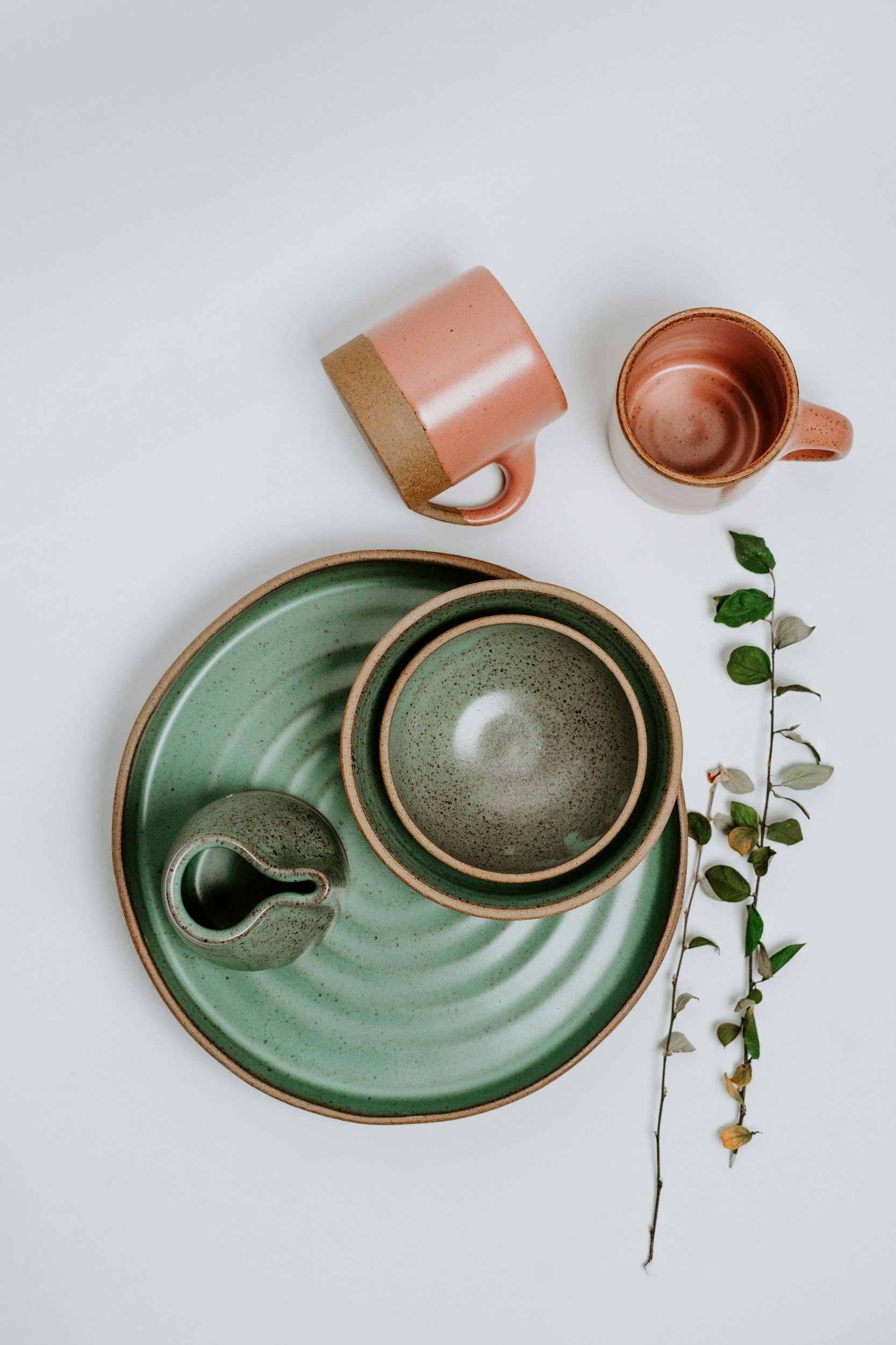 Arrangement of green and brown ceramic dishes and cups on a white surface, with two pink ceramic mugs and a small green leafy branch.