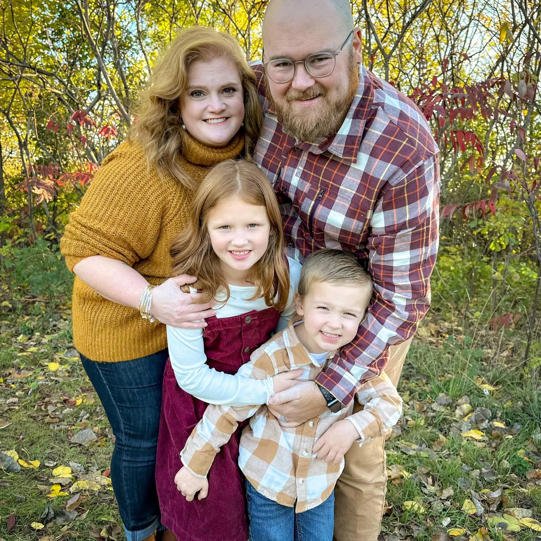 A family of five posing outdoors among autumn foliage, smiling. The woman on the left has red hair and is wearing a mustard yellow sweater, the man on the right has a beard, glasses, and is wearing a plaid shirt. Two young girls and a boy, all with light-colored hair, are in front of the parents, close together, smiling at the camera. The ground is covered with fallen leaves, and the background features trees with colorful fall leaves.