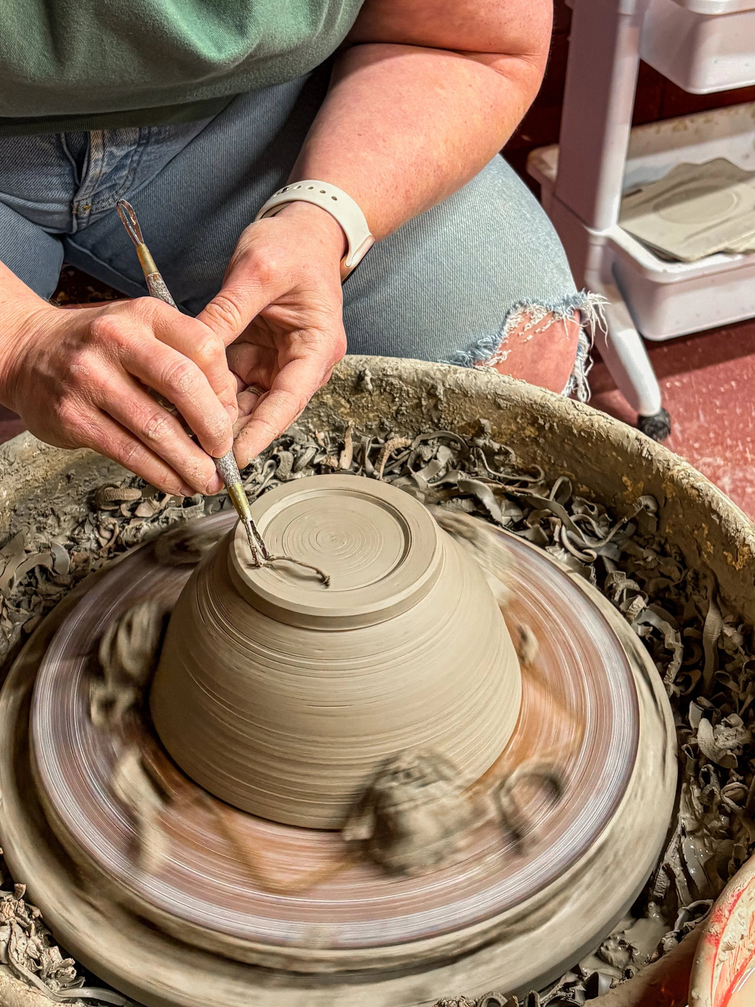 A person in green shirt and blue jeans is shaping a clay vessel on a pottery wheel, with scraps of clay around.