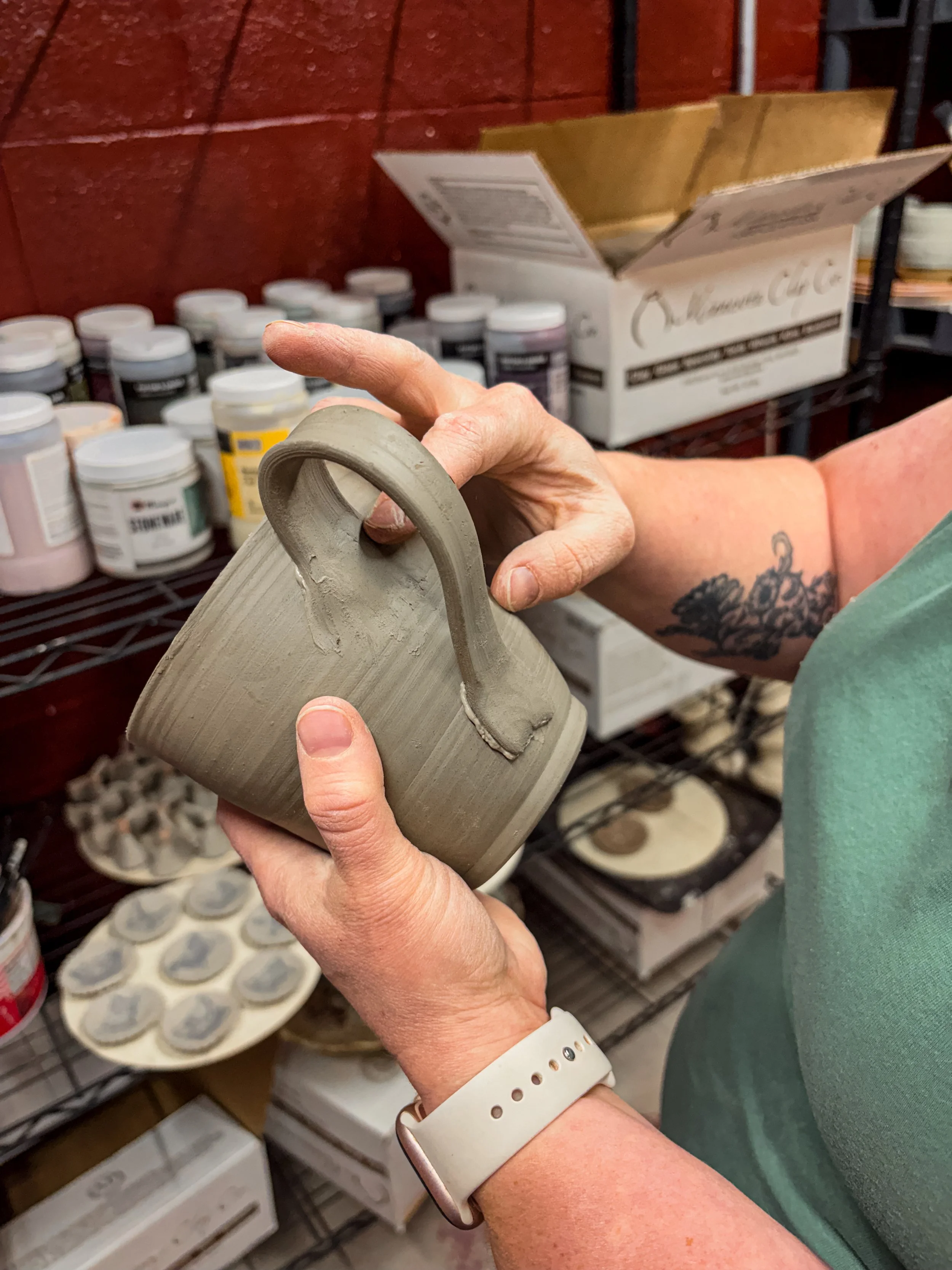 Person holding a gray ceramic mug in a pottery studio with shelves of paint jars and a box of glazed ceramics in the background.