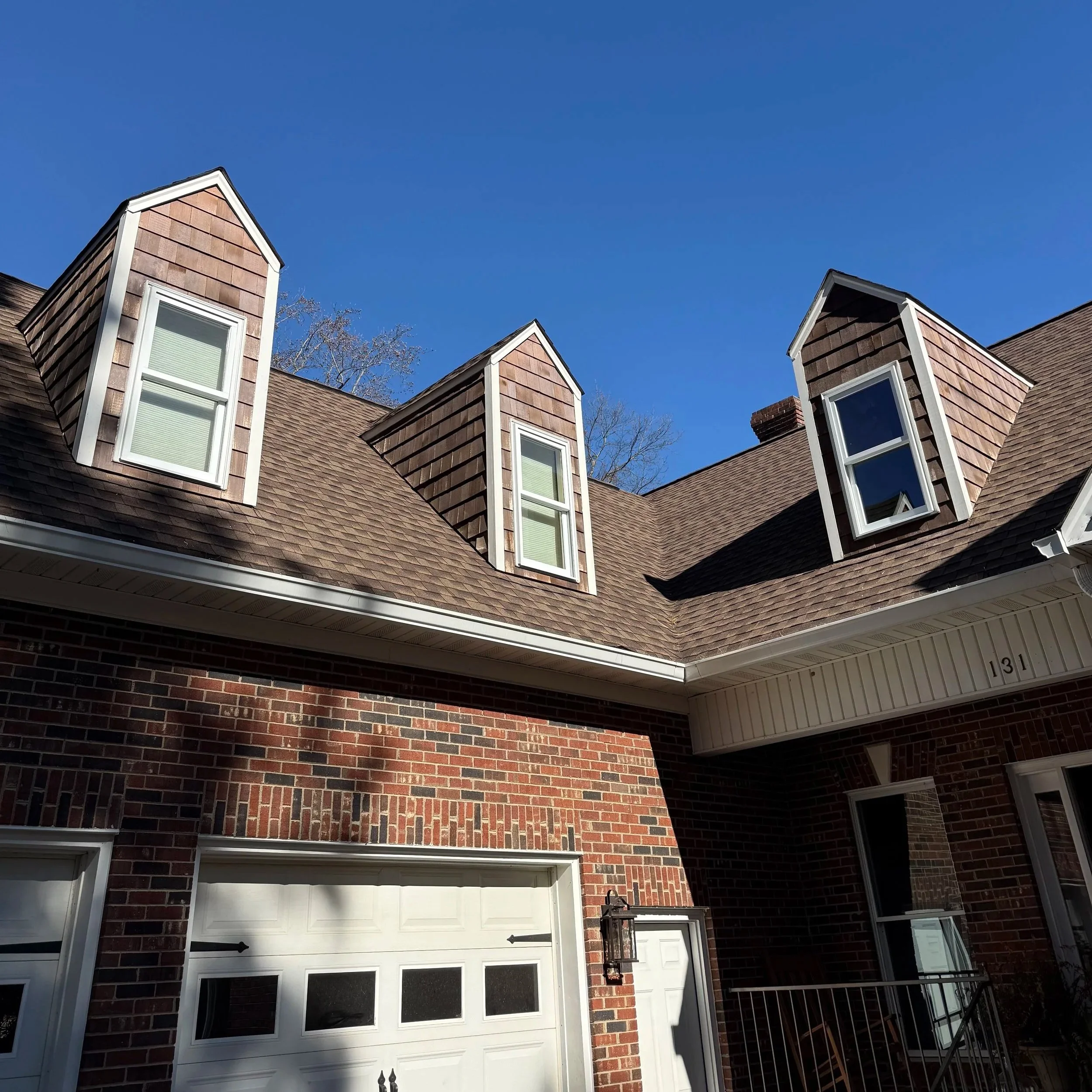 The image shows the upper part of a brick house with a brown shingled roof and multiple dormer windows against a clear blue sky.