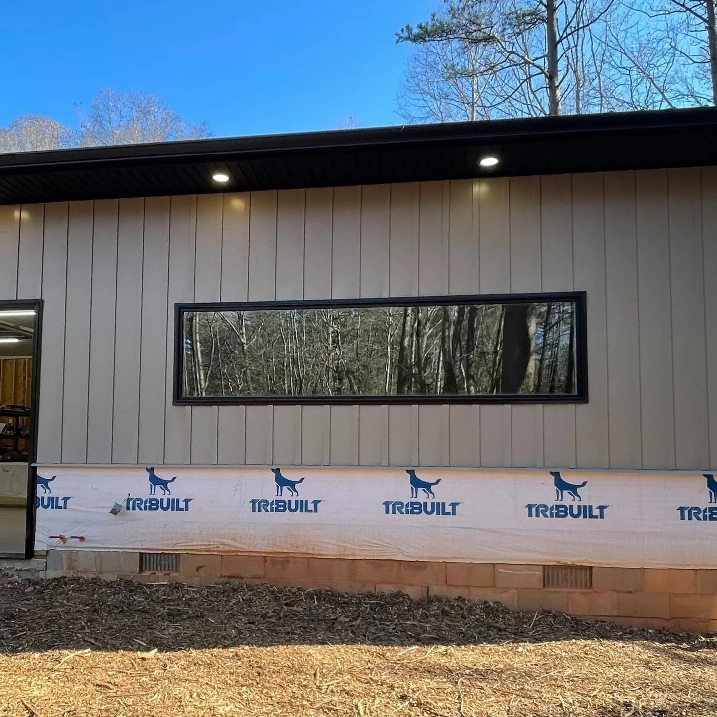 Exterior of a house under construction with a beige vertical siding, a long rectangular window, and a partly finished lower wall with Tyvek Tribuilt house wrap. The sky is blue with some trees in the background.