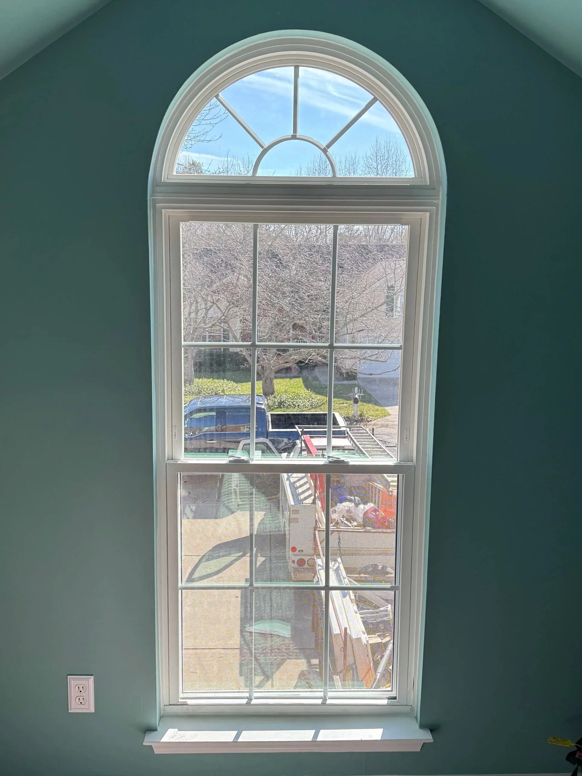 View of a tall, arched window with divided panes, looking out onto a driveway with parked vehicle and a yard with trees, under a clear blue sky.