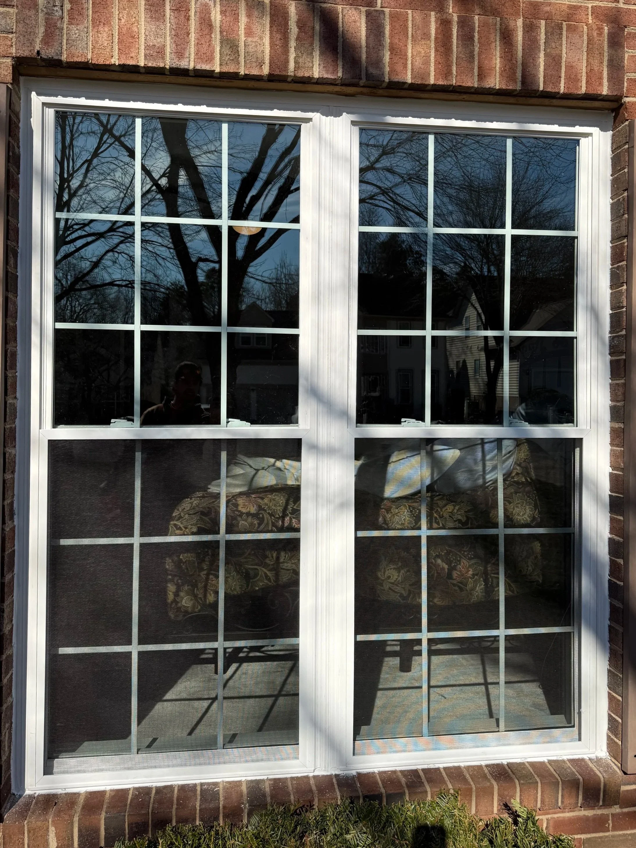 A sliding glass door with white grid frames on a brick house wall, reflecting trees and the sky, with a patterned curtain inside and a person faintly visible through the glass.